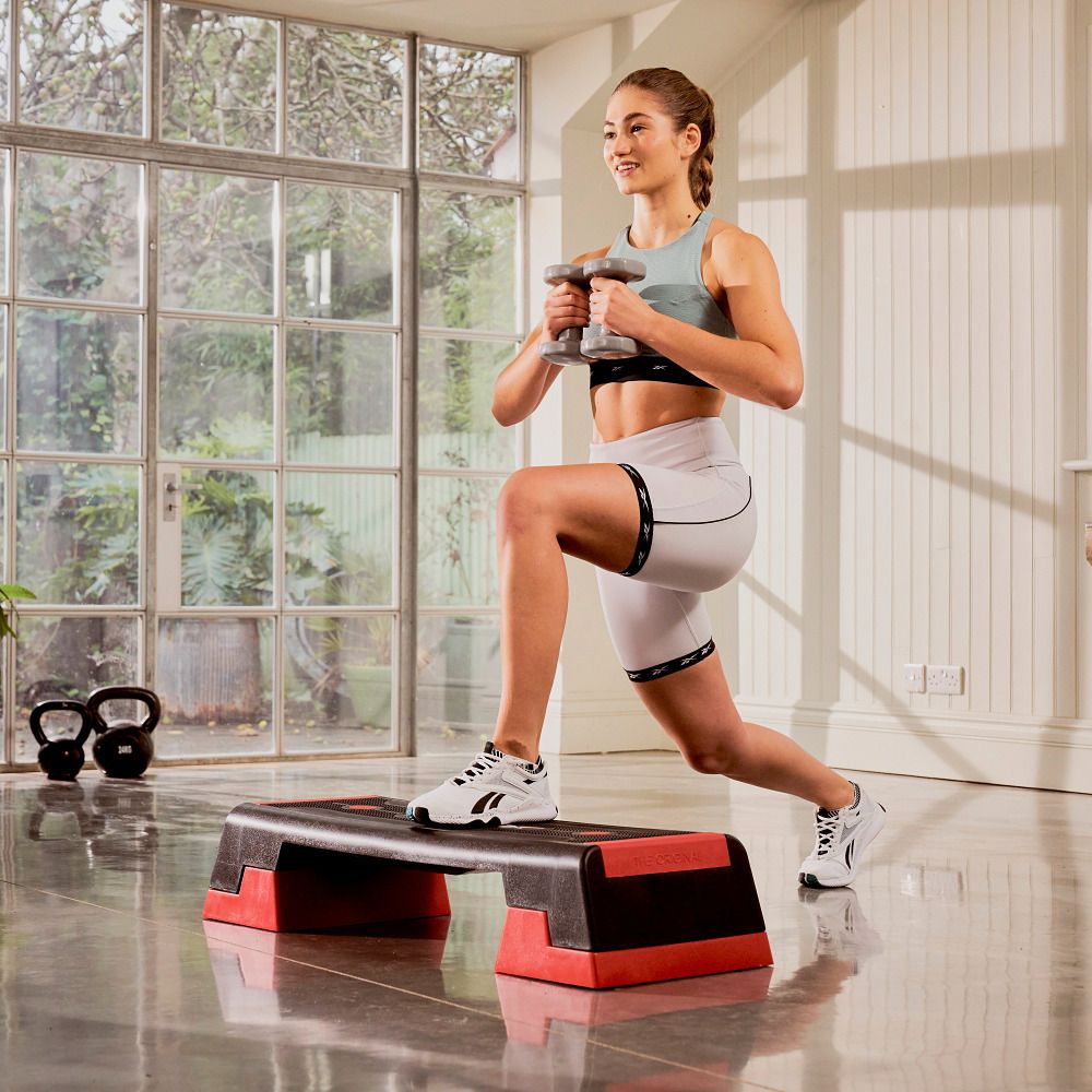 Schwarzer und roter Reebok Step. Eine Frau macht eine Ausfallschrittübung mit Hanteln. Im Hintergrund sind Kettlebells zu sehen.