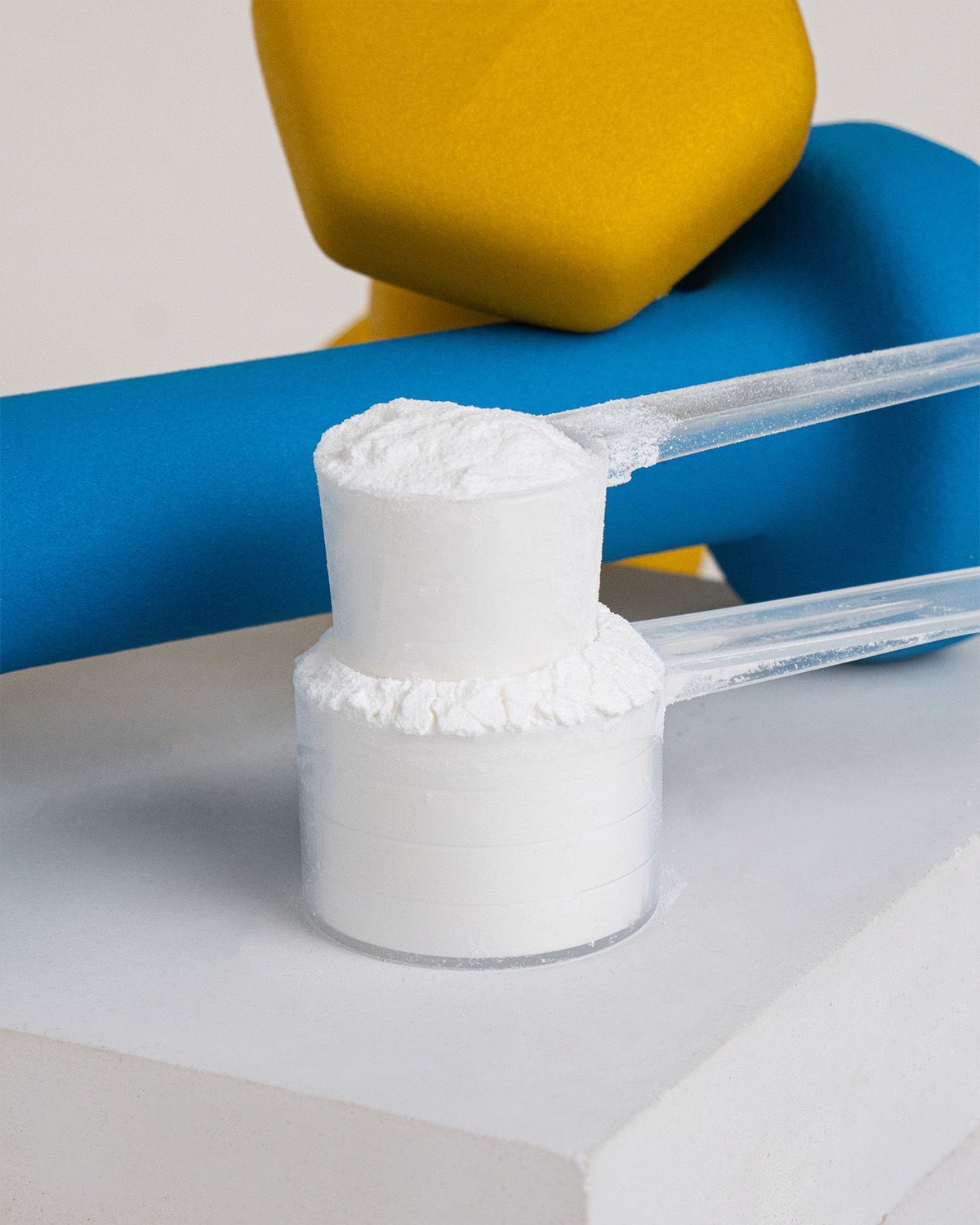 Ein Messlöffel gefüllt mit weißem BCAA-Pulver, neben einem blauen und gelben Hantel.