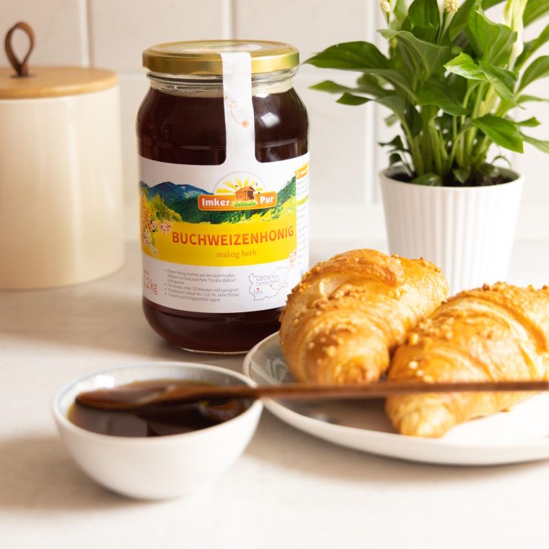 Honigglas mit Etikett neben Croissants und einer Schale Honig. Aufschrift: ImkerPur Buchweizenhonig. Goldener Deckel. Honigfarbe.