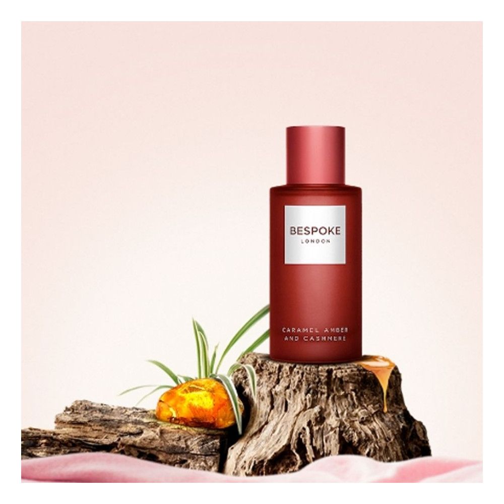 Rote Parfumflasche auf Holzstamm. Aufschrift: Bespoke London, Karamell Amber & Kaschmir. Tropfen von Flüssigkeit.