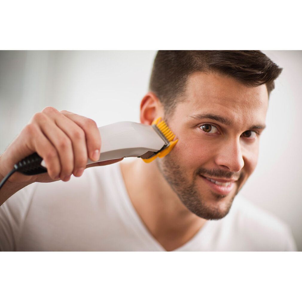 Mann rasiert sich mit einem weißen Haarschneider. Gelber Aufsatz. Lächelt.