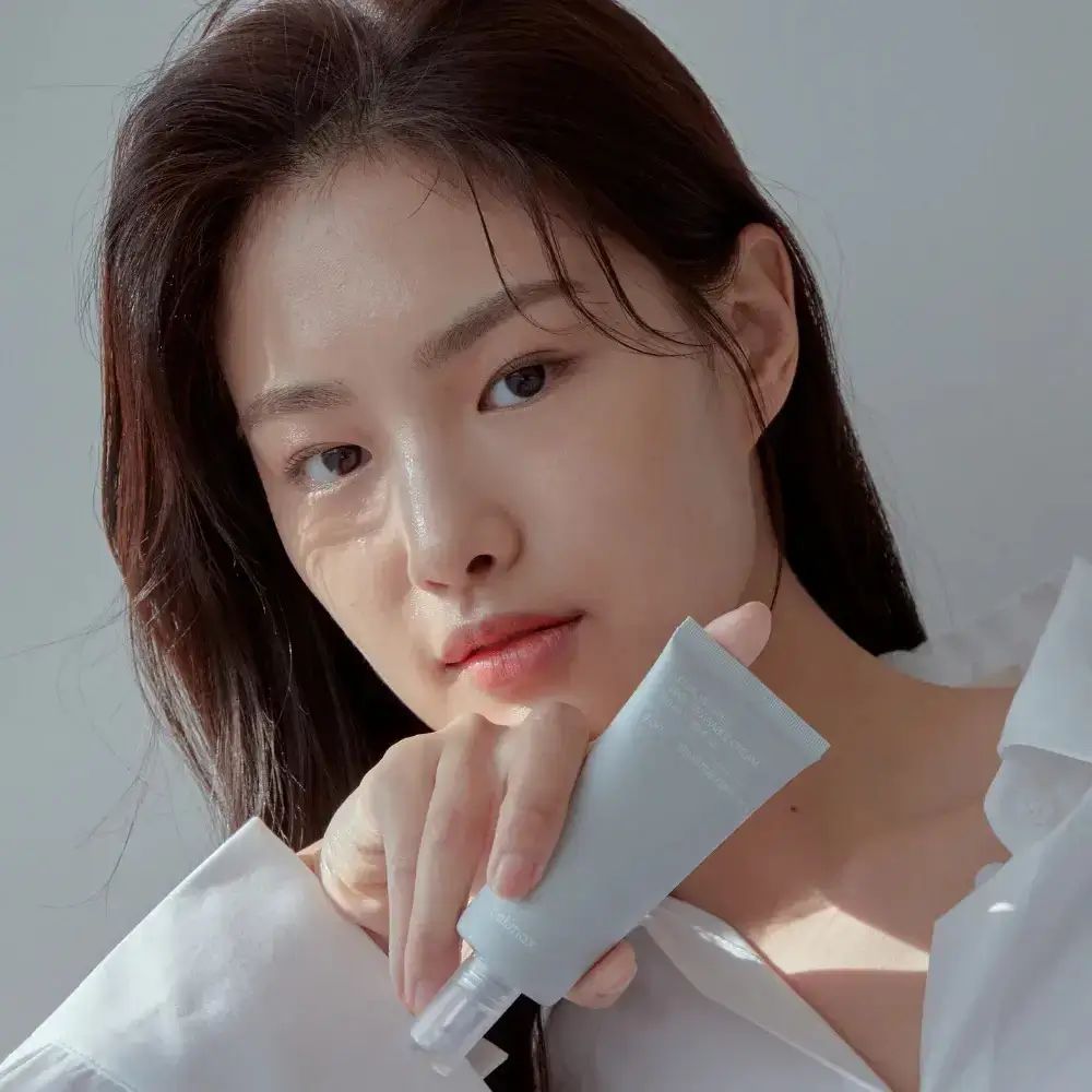 Frau hält eine graue Tube. Aufschrift nicht lesbar. Sie trägt ein weißes Hemd. Natürliches Licht.