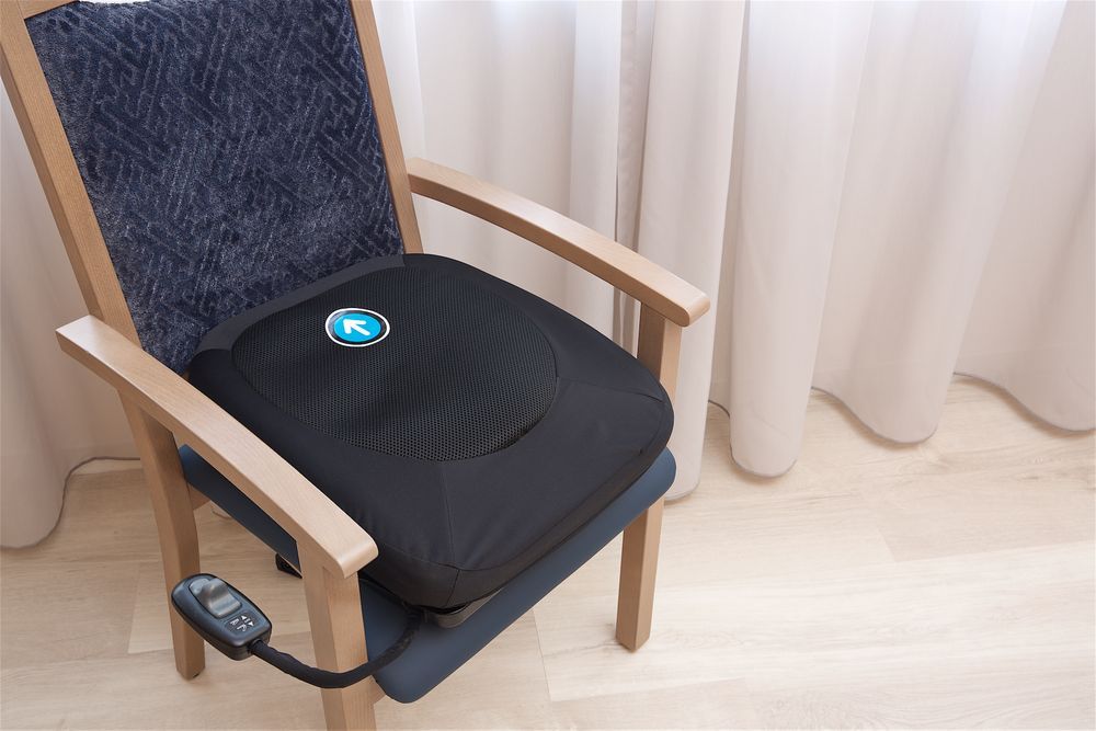 Schwarze elektrische Aufstehhilfe auf einem Stuhl. Blaues Logo sichtbar. Fernbedienung.
