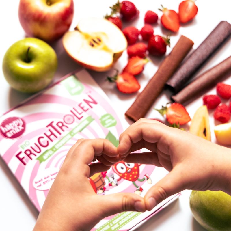 Hände formen Herz über der Verpackung von BIO Fruchtrollen. Früchte liegen daneben.