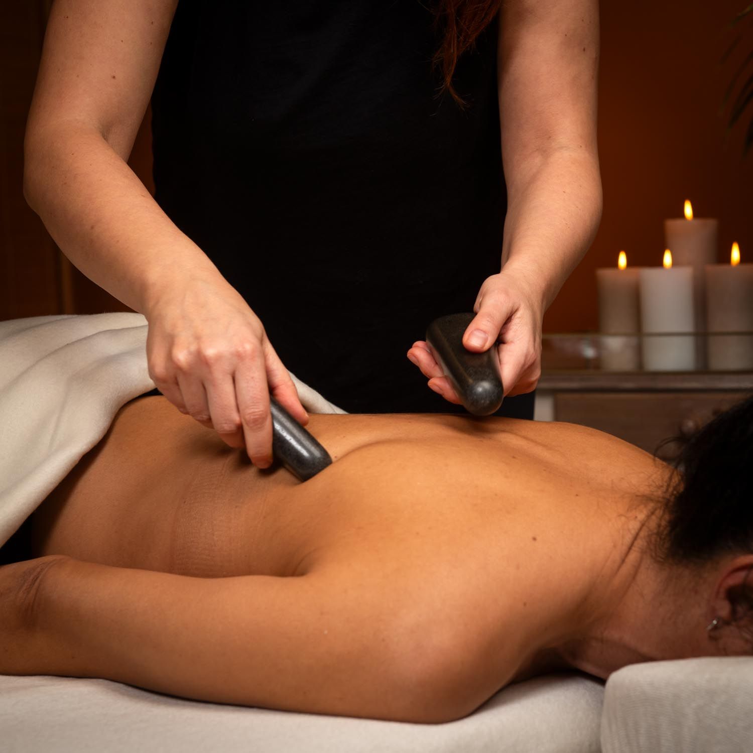 Person hält zwei dunkle, längliche Steine auf dem Rücken einer Person. Im Hintergrund Kerzen.