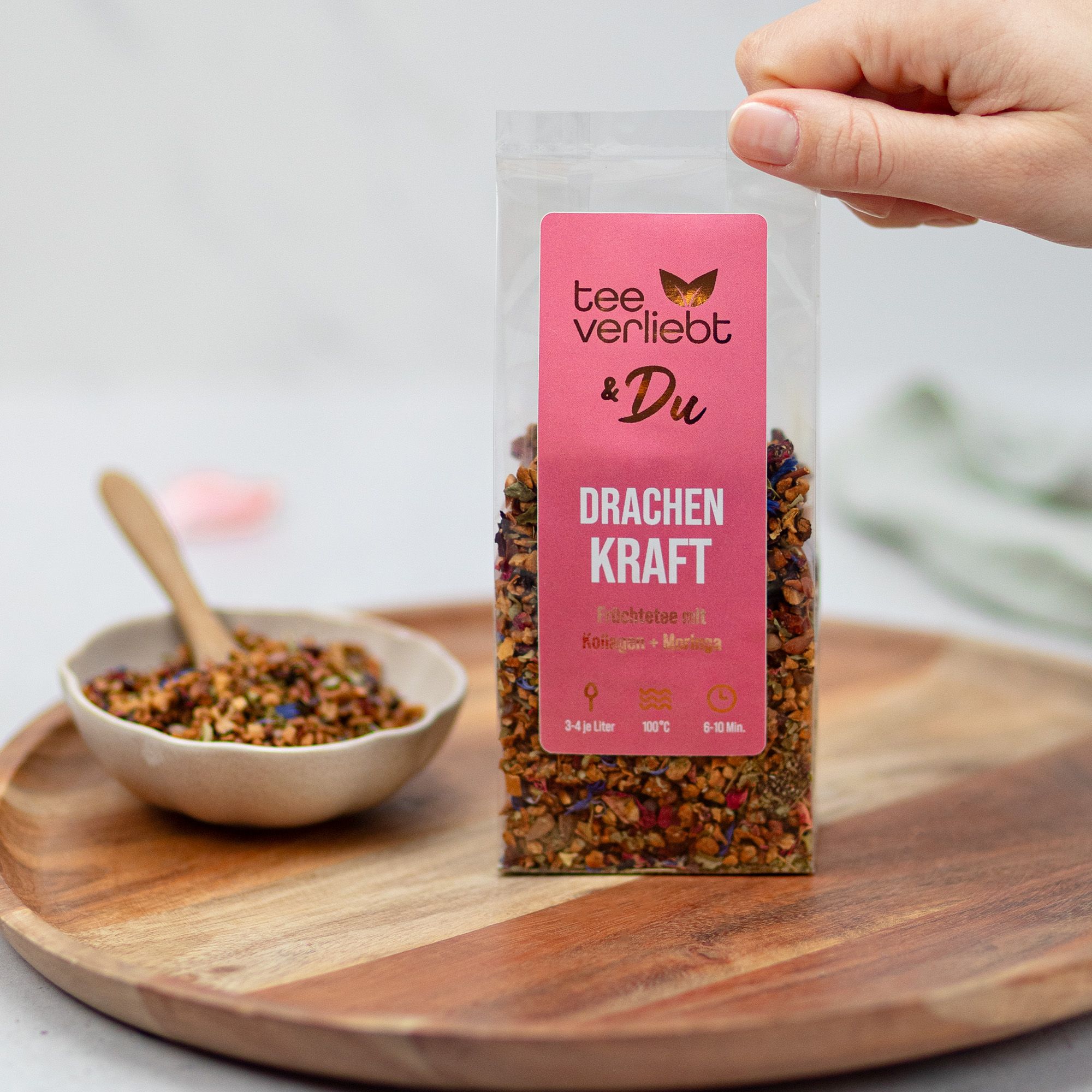 Früchtetee in transparenter Verpackung, gehalten von einer Hand. Auf einem Holztablett, daneben eine Schale mit Tee.