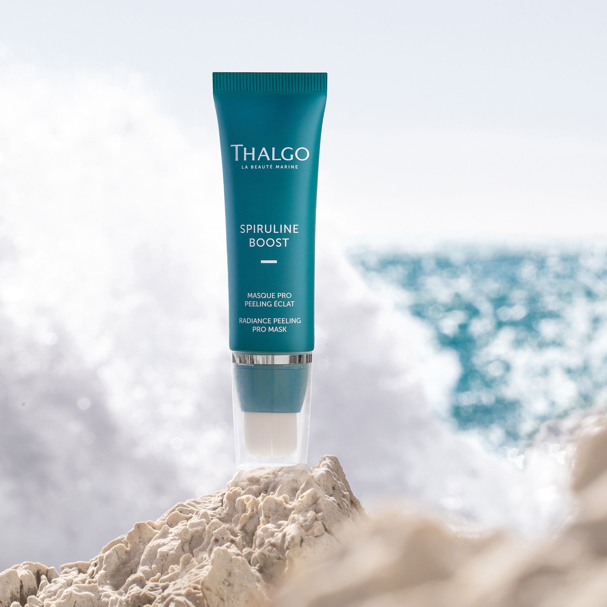 Türkisfarbene Tube mit weißem Pinsel. Aufschrift: THALGO, SPIRULINE BOOST, MASQUE PRO PEELING ÉCLAT, RADIANCE PEELING PRO MASK. Vor Meereskulisse.