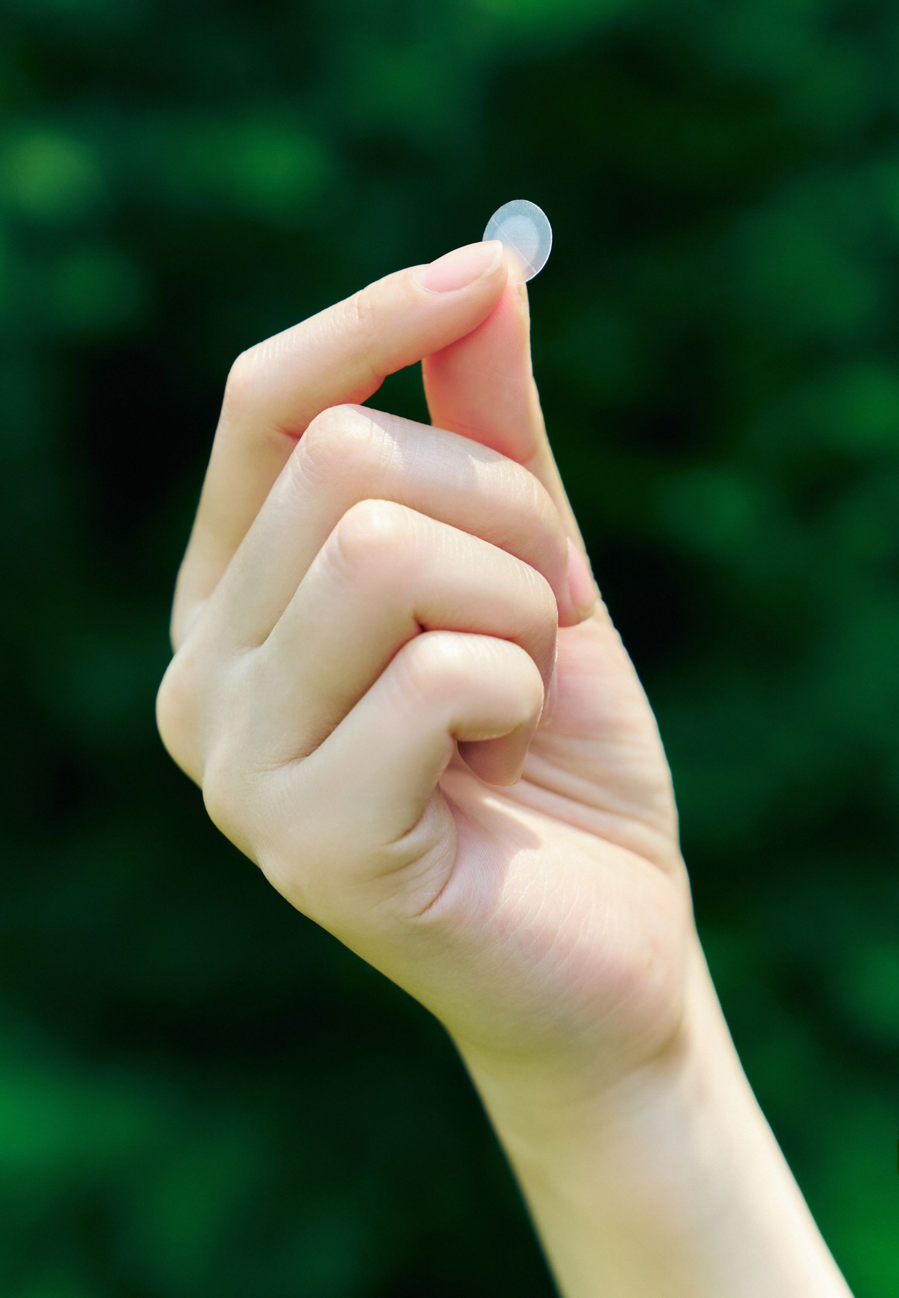 Hand hält einen transparenten, runden Patch. Grüner Hintergrund.
