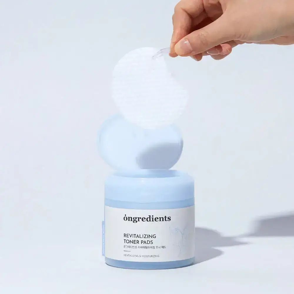 Geöffnete Dose mit Pads, Hand entnimmt Pad. Aufschrift: "ongredients Revitalizing Toner Pads". Blauer Hintergrund.