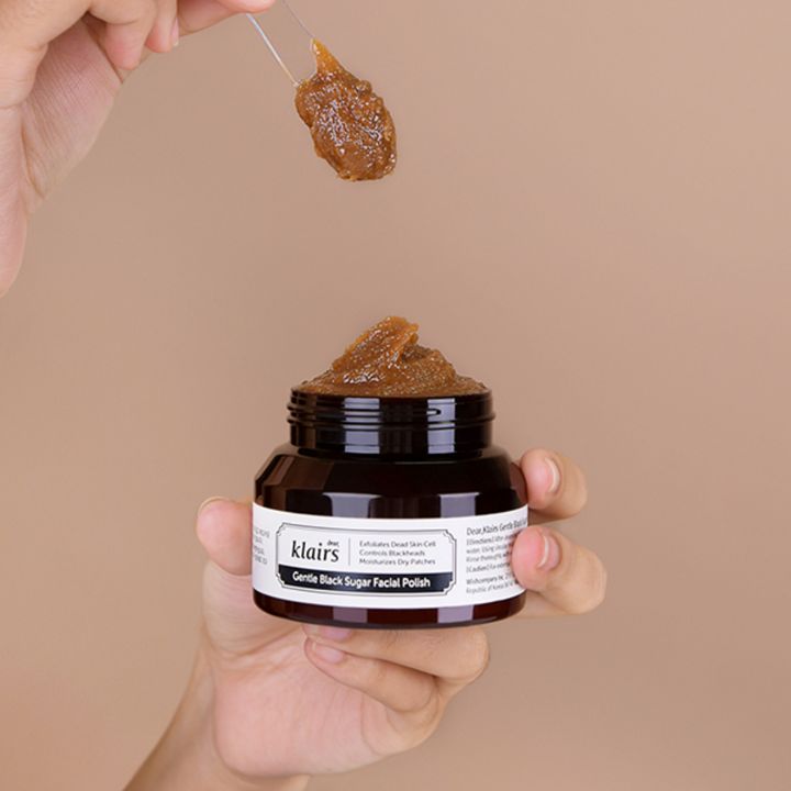 Glas mit brauner Creme, gehalten von Händen. Löffel mit Creme darüber. Aufschrift: dear, klairs Gentle Black Sugar Facial Polish.