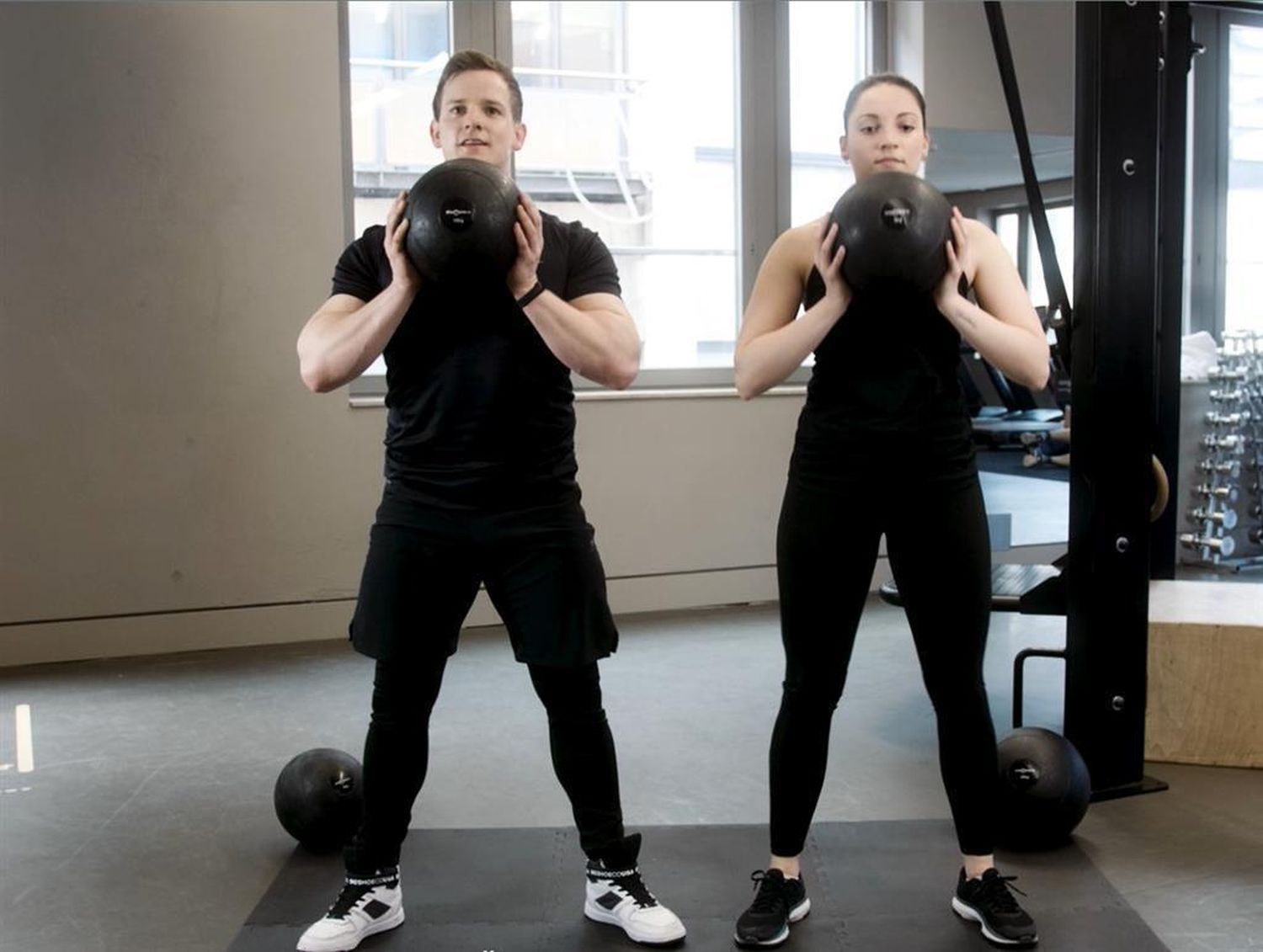 Zwei Personen in Sportkleidung stehen und halten Slambälle. Fitnessstudio-Umgebung.