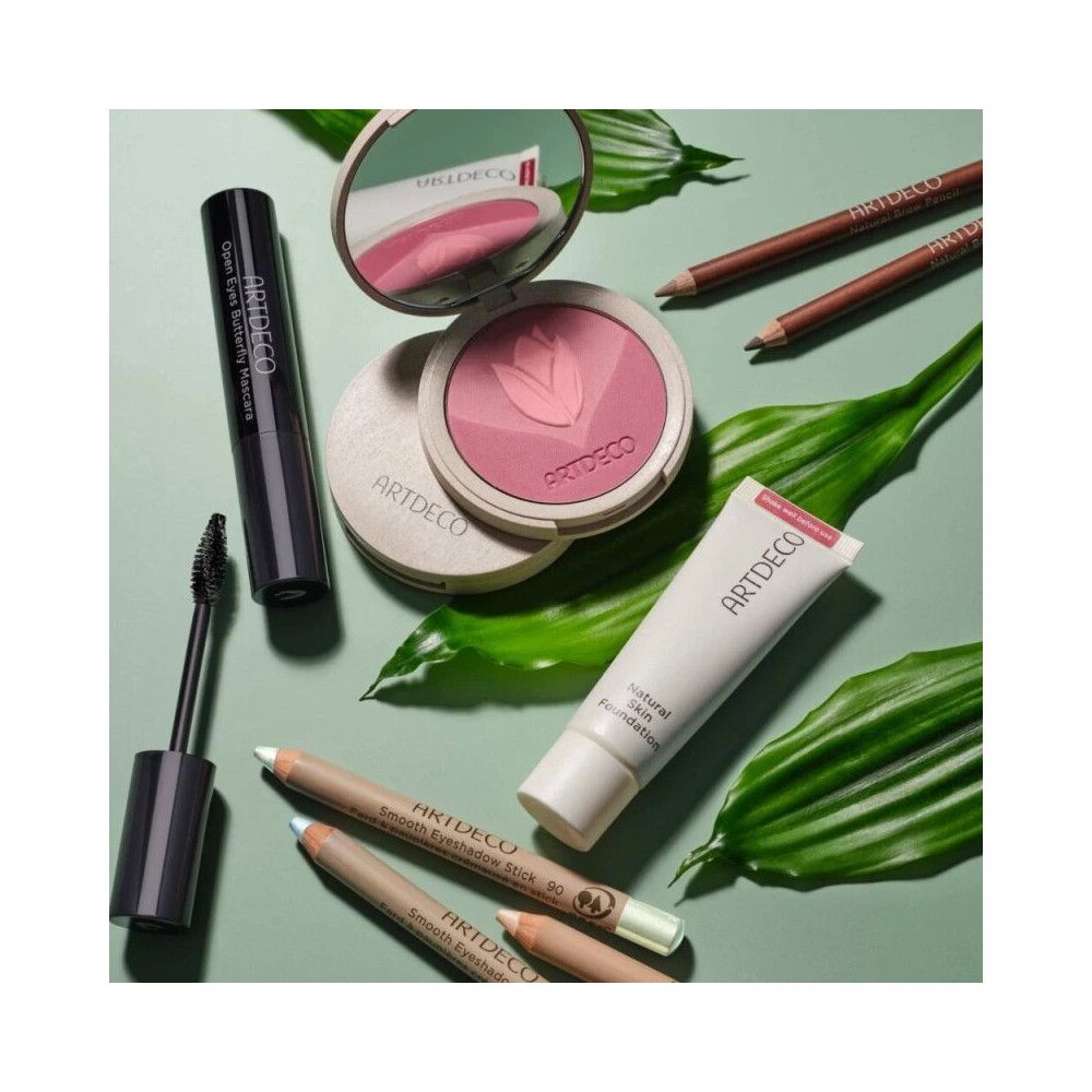 Kosmetikprodukte auf grünem Hintergrund. Enthalten: Mascara, Puder, Foundation, Augenbrauenstifte. Artdeco-Logo sichtbar.