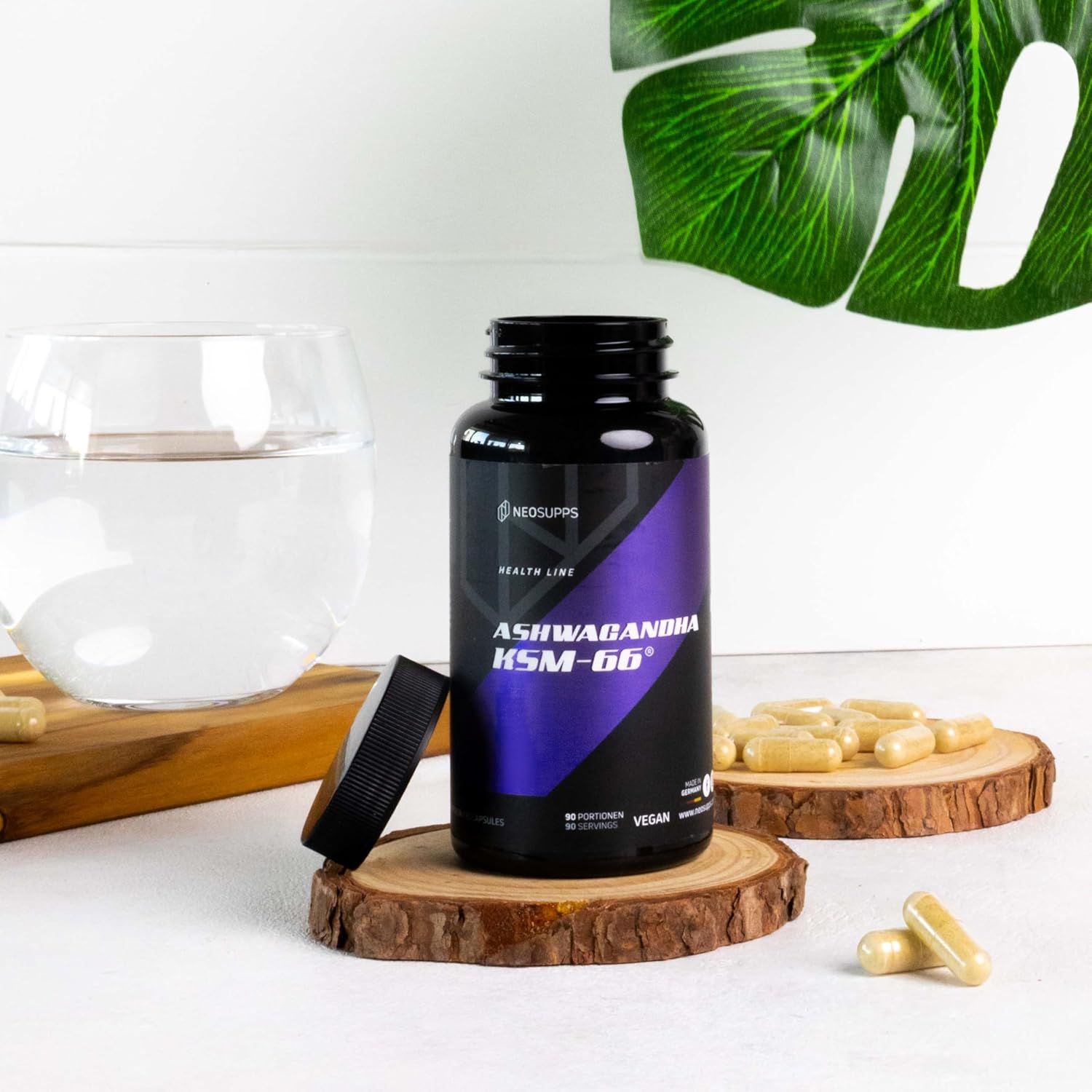 Schwarze Flasche "ASHWAGANDHA KSM-66" mit geöffnetem Deckel, Kapseln und Glas Wasser auf Holzscheiben. Marke: Neosupps.