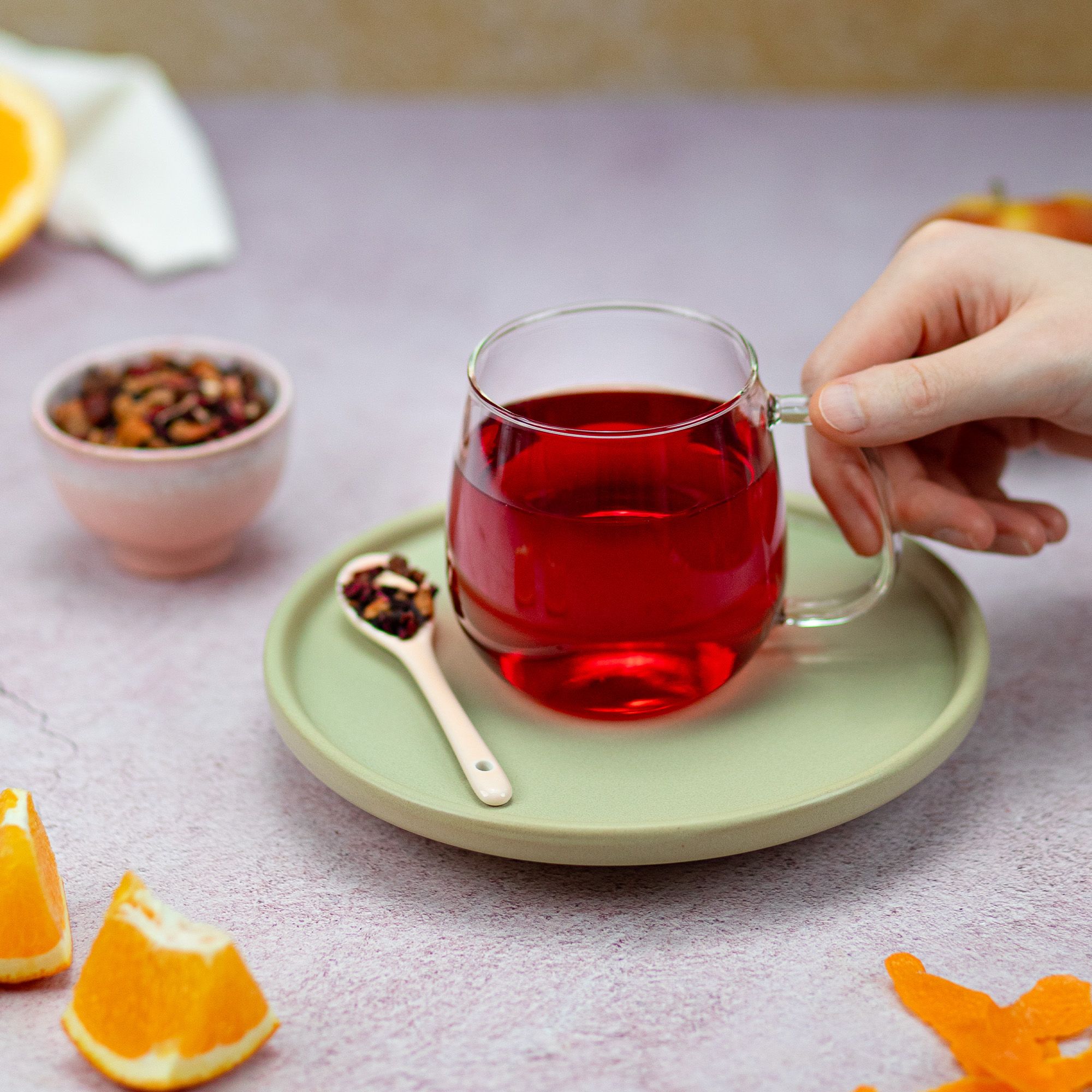 Glasbecher mit rotem Tee auf Untertasse. Hand hält Becher. Löffel mit Tee daneben. Orangenstücke.
