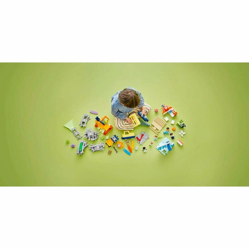 Kind sitzt auf grünem Hintergrund, umgeben von LEGO Duplo-Teilen. Gelber Zug und Zubehör.