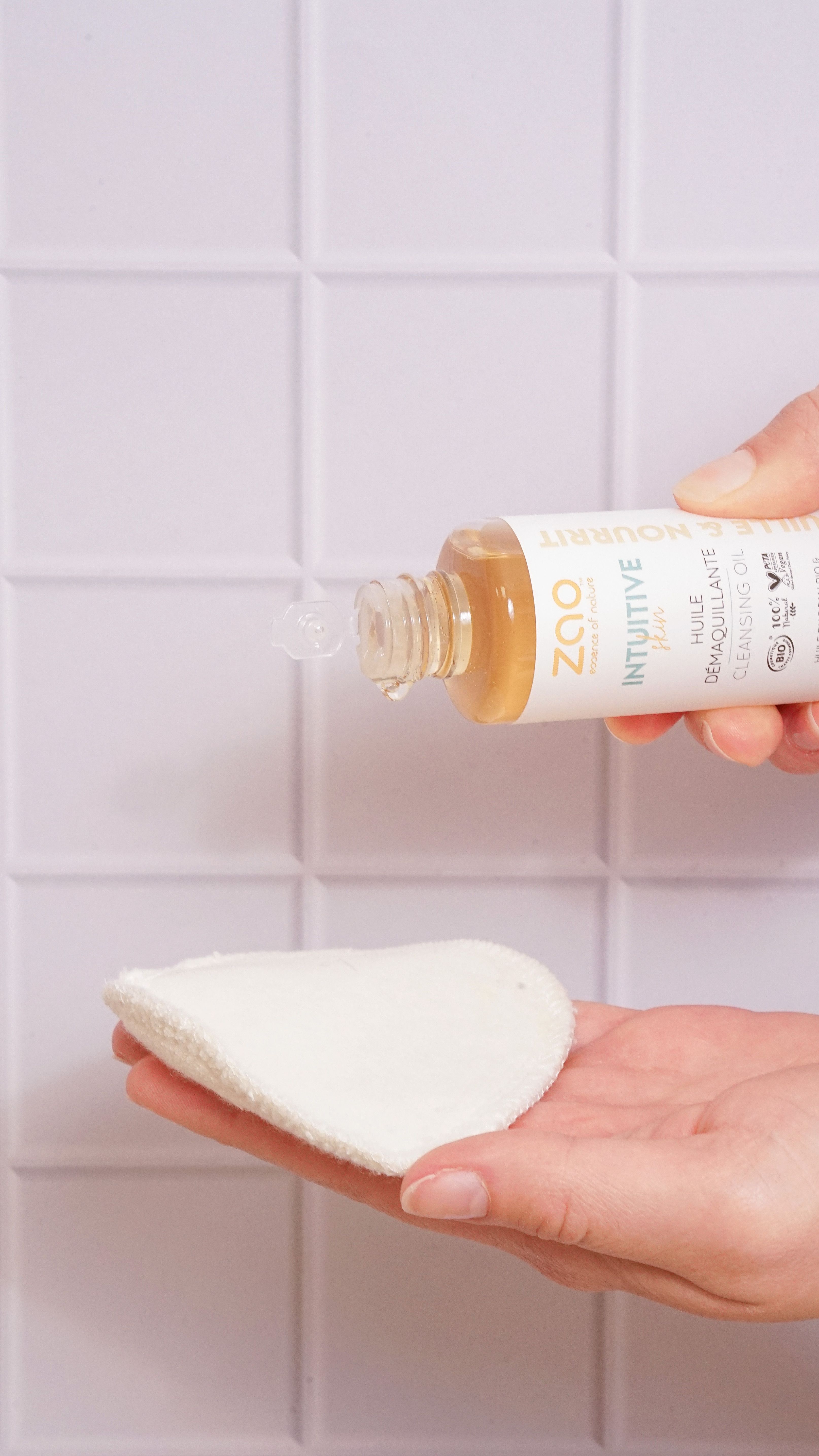 Helle Flüssigkeit tropft aus einer Flasche auf ein Wattepad. Flasche mit Holzverschluss, Etikett mit Text. Hintergrund: weiße Fliesen.