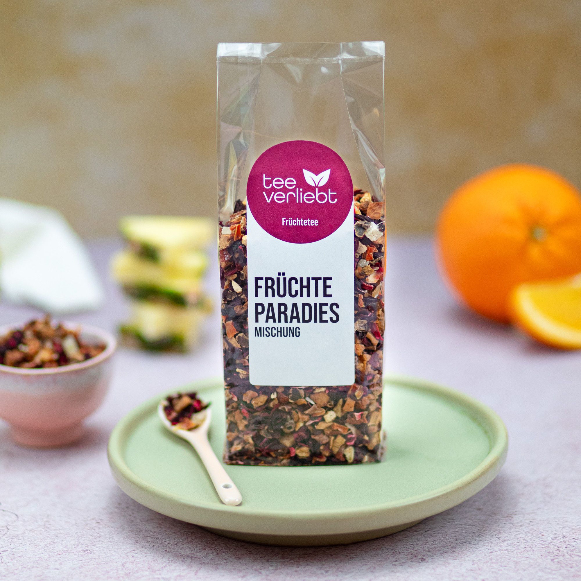 Transparente Verpackung mit loser Früchtetee-Mischung. Auf Teller mit Löffel und Obst. Logo und Produktbezeichnung: Früchte Paradies Mischung.