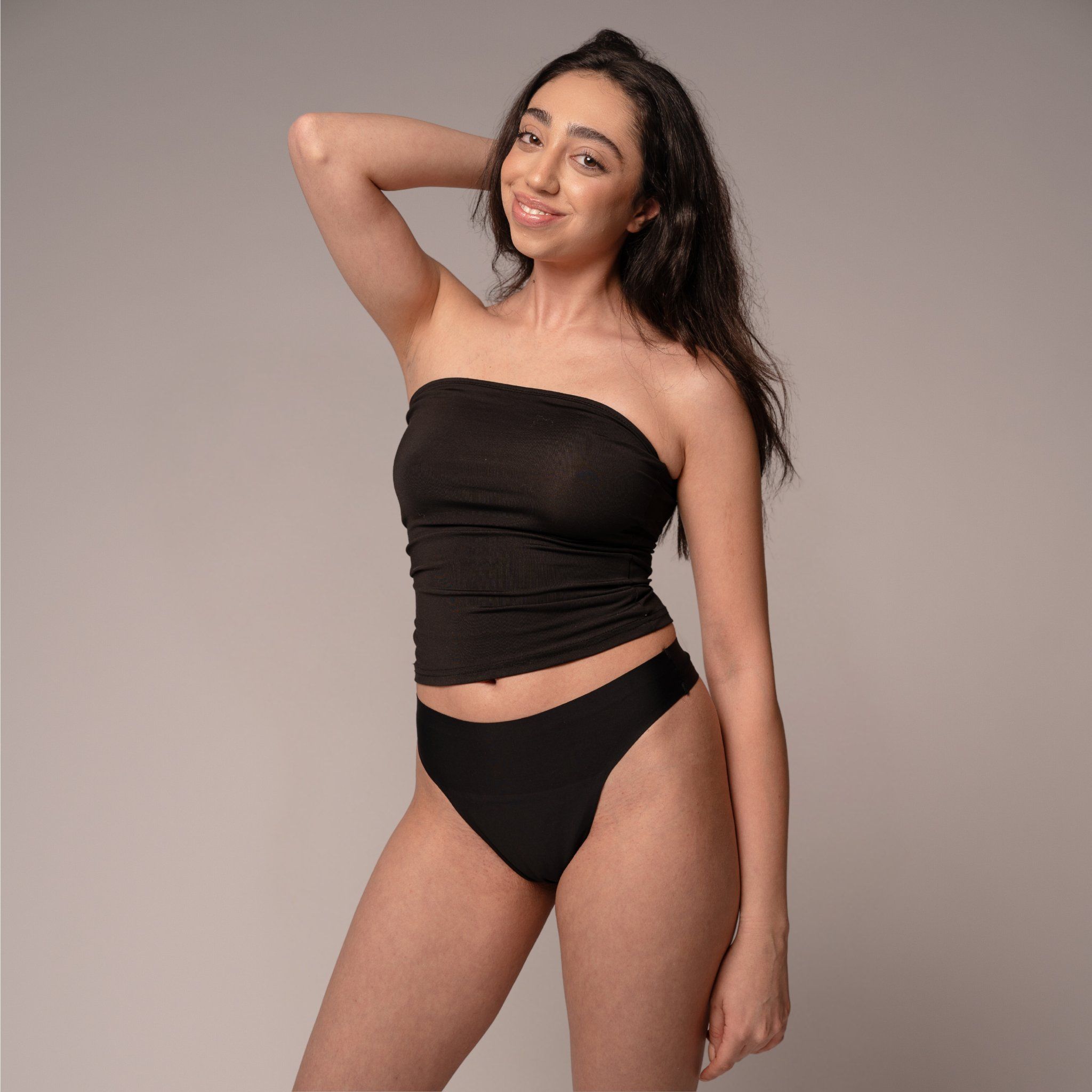 Eine Frau in einem schwarzen Tanktop und einem schwarzen Slip. Sie lächelt und berührt ihre Haare.