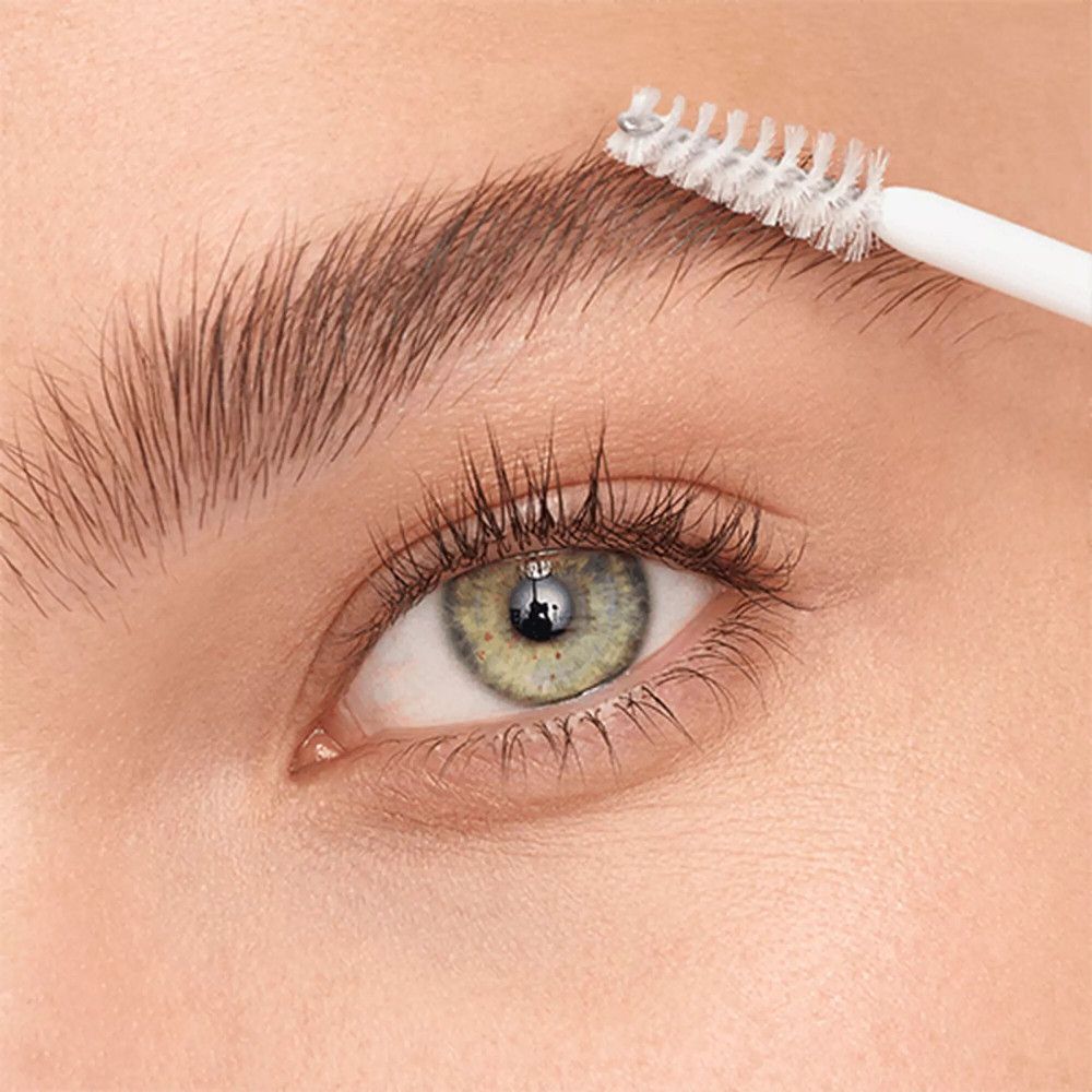 Auge mit Augenbraue, die mit einer Bürste gekämmt wird. Helle Haut, grüne Augen.