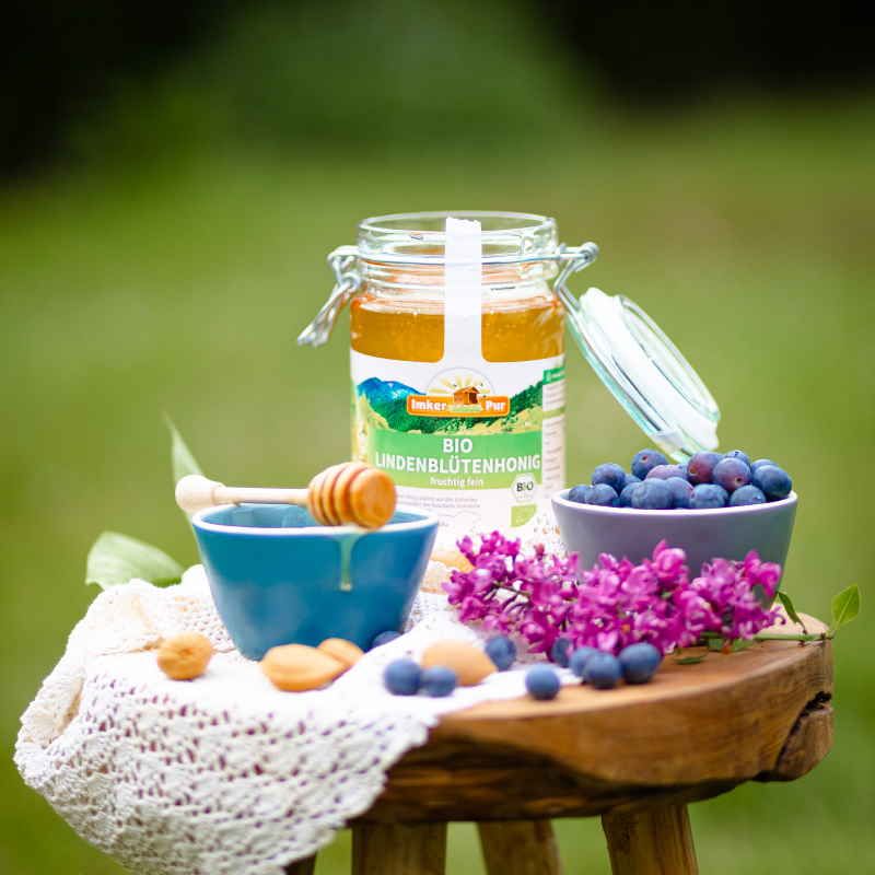 Honigglas "ImkerPur Bio Lindenblütenhonig" auf Holzschemel. Honig, Blaubeeren, Blüten und Löffel.