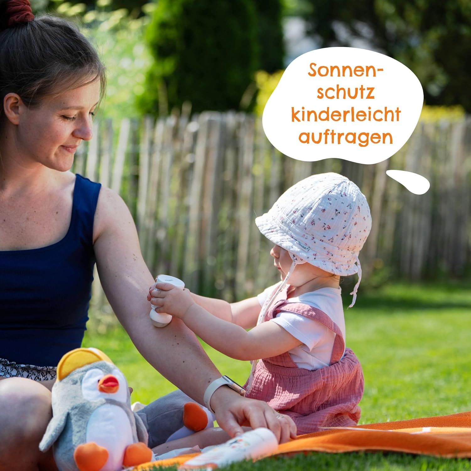 Mutter trägt Sonnenroller auf Arm eines Kindes auf. Auf Rasen sitzend. Sprechblase: Sonnenschutz kinderleicht auftragen. Kuscheltier Pinguin.