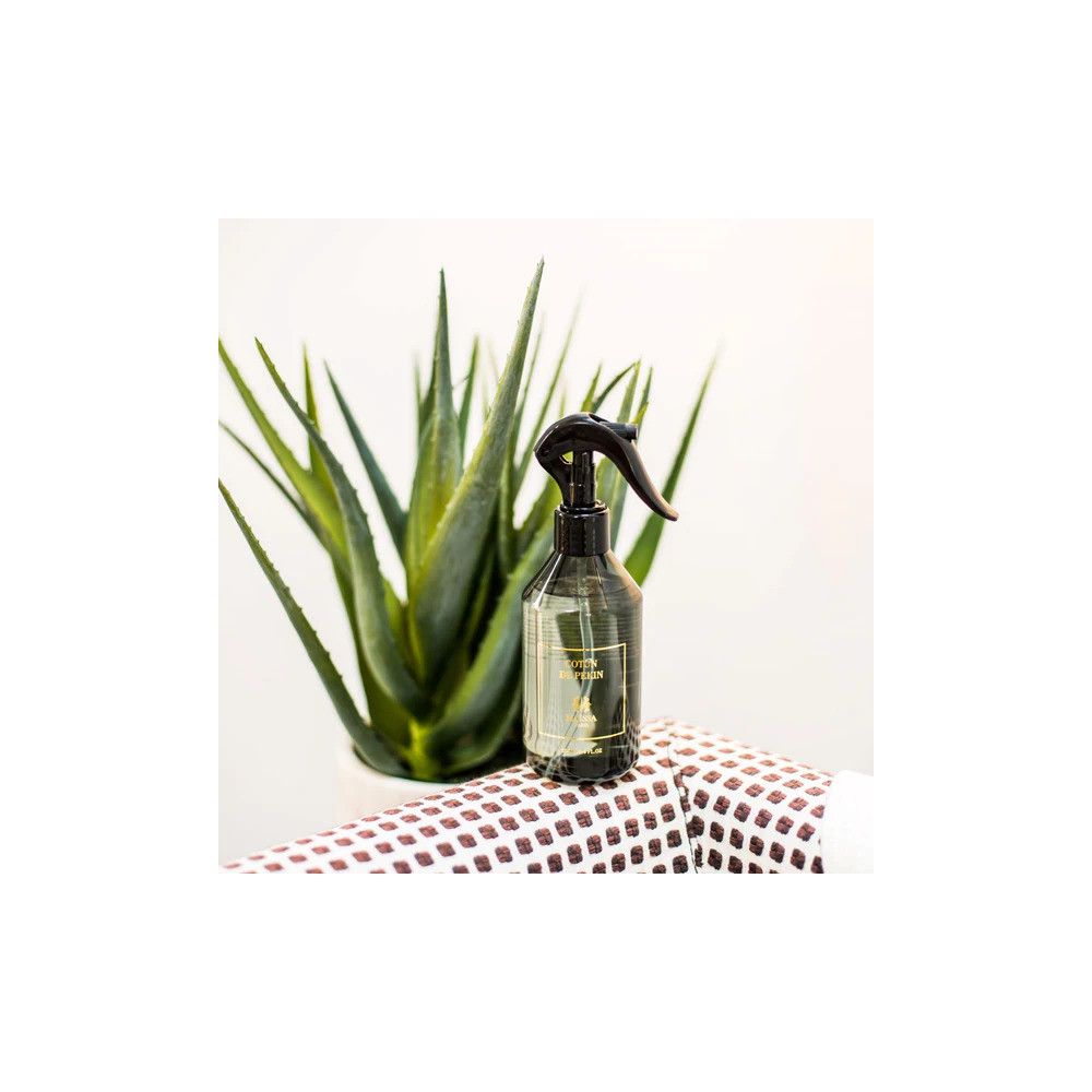 Sprühflasche neben einer Pflanze auf gemustertem Stoff. Flasche mit schwarzem Sprühkopf und Etikett.