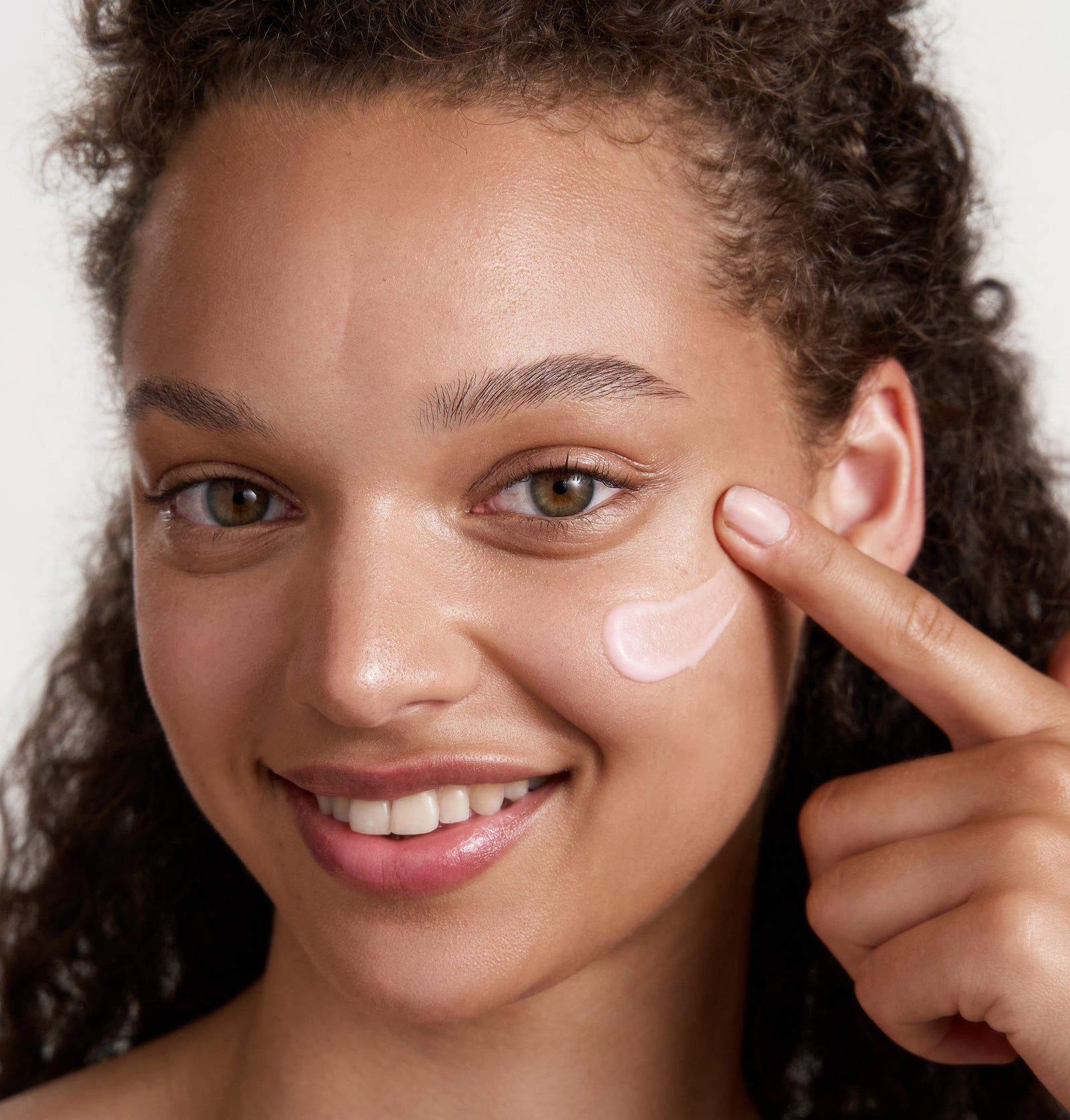 Frau mit lockigem Haar, die Creme auf die Wange aufträgt. Rosa Creme auf der Haut sichtbar.