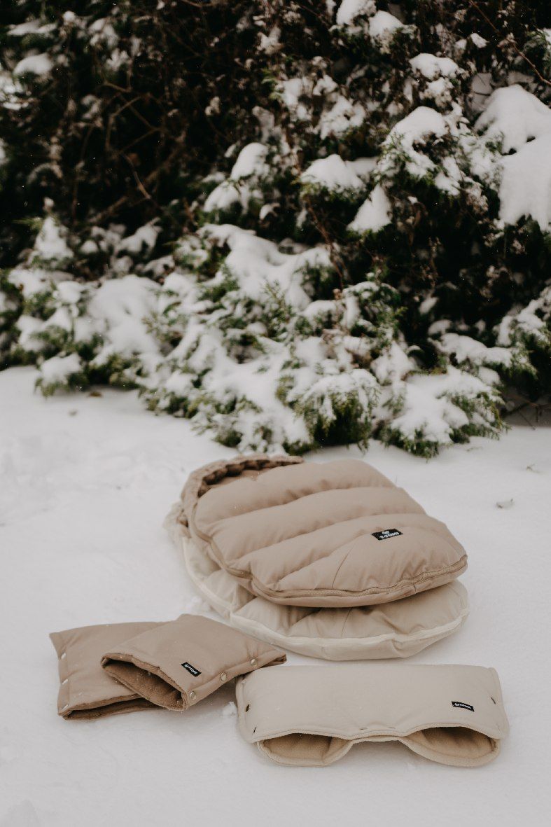 Gepolsterter Babyschlafsack in Beige mit passenden Handwärmern und einer Unterlage im Schnee.