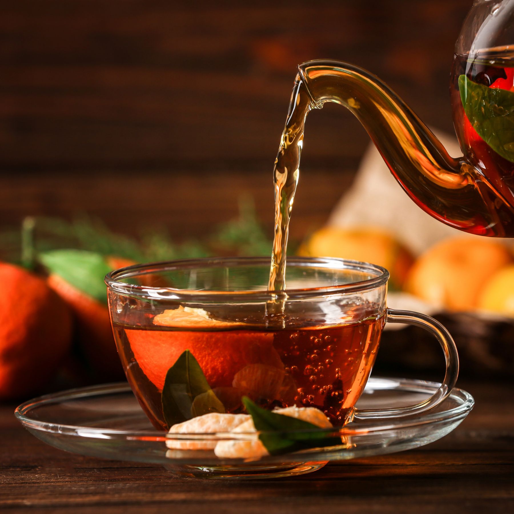 Tee wird aus einer Glaskanne in eine klare Tasse gegossen. Auf einem Unterteller liegen Kekse und Blätter.