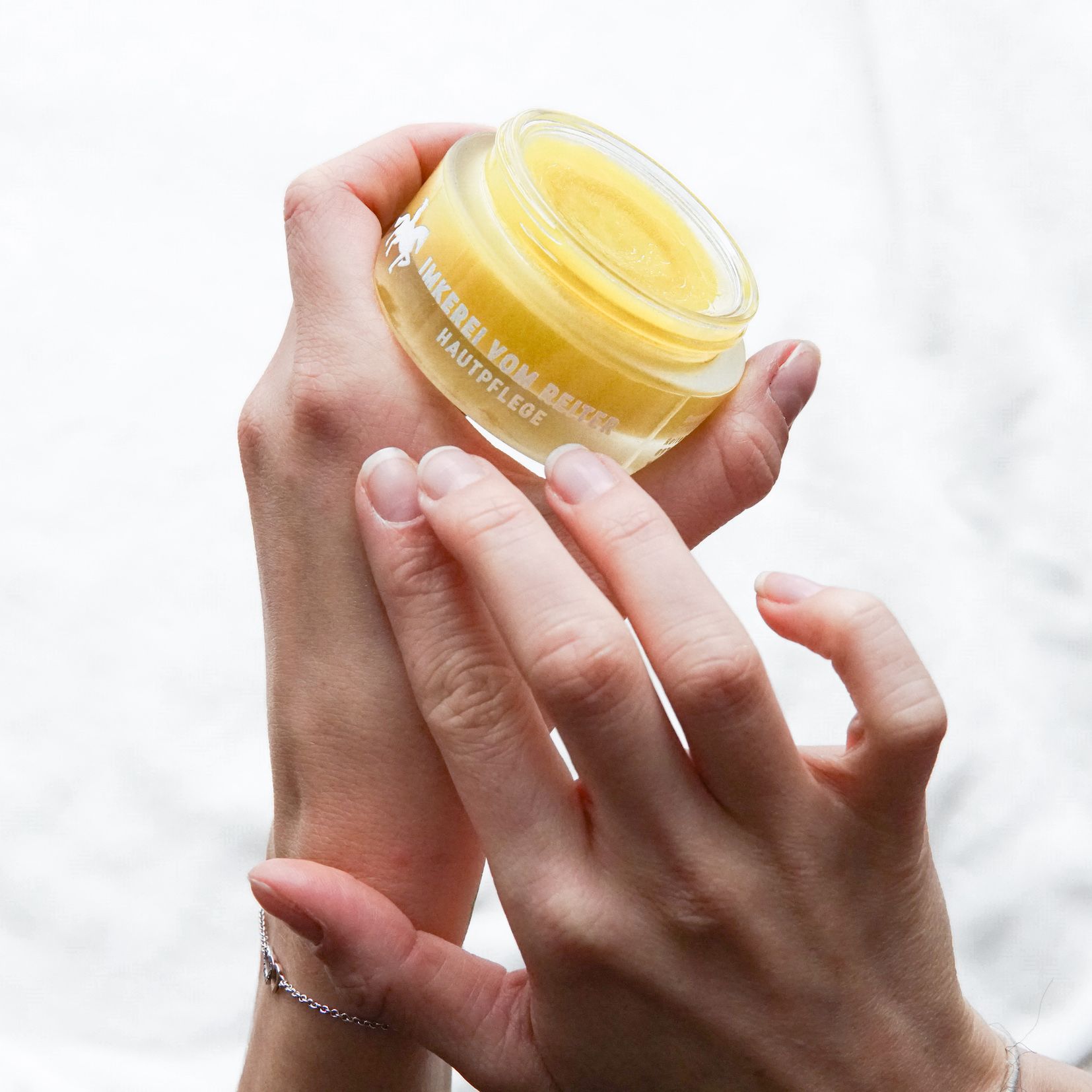 Hände halten einen geöffneten Creme-Tiegel. Aufschrift: Imkerei vom Reiter Hautpflege. Gelbe Creme sichtbar.