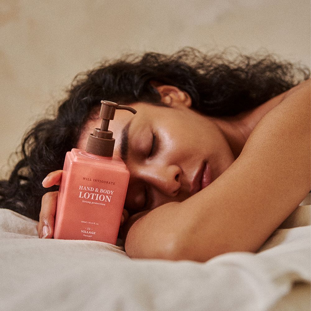 Person hält eine rosa Lotion-Flasche. Aufschrift: Hand & Body Lotion, Strong protection. Marke: Village 11 Factory. Person liegt auf Bett.