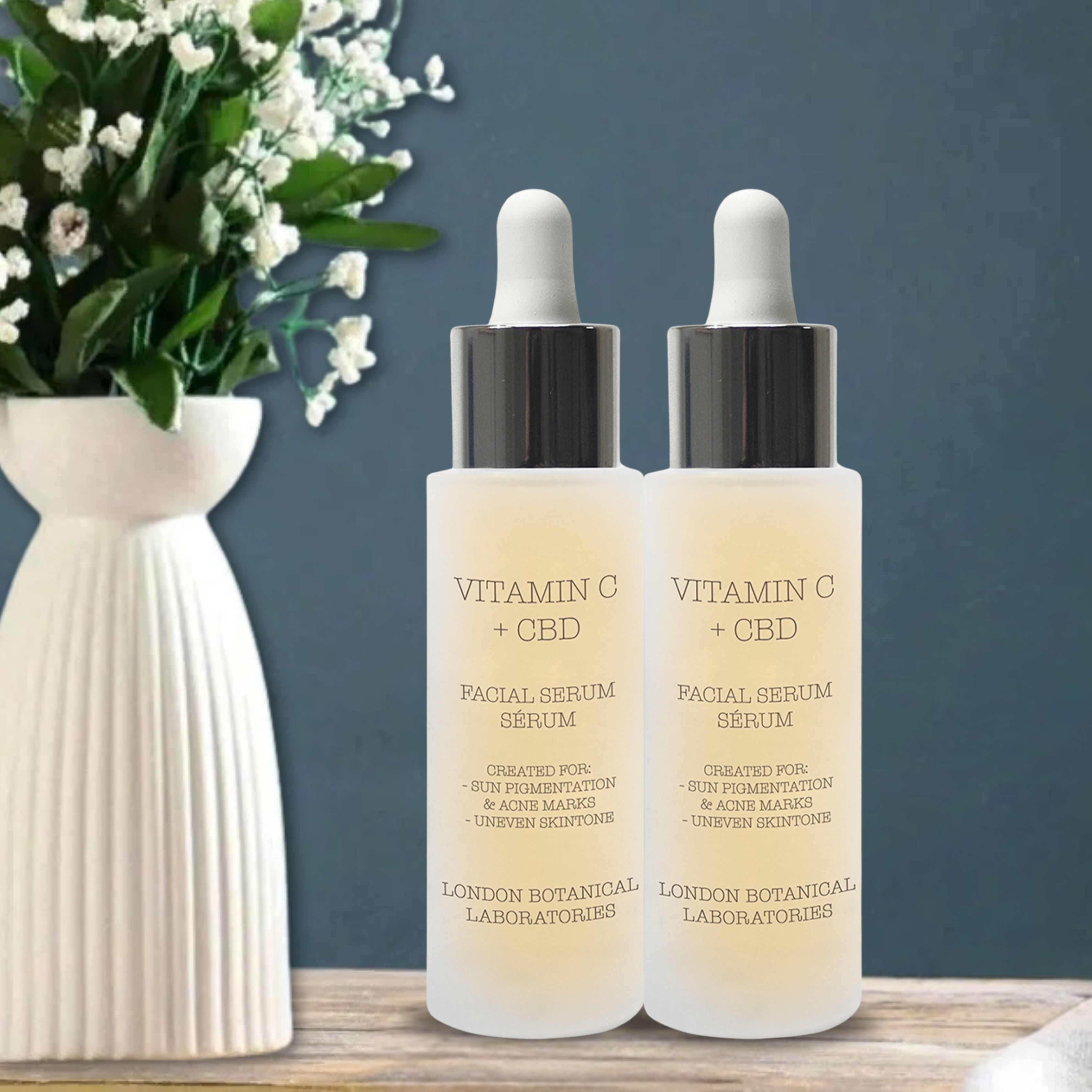 Zwei Flaschen Serum mit Pipetten. Aufschrift: Vitamin C + CBD. Marke: London Botanical Laboratories. Hintergrund: Vase mit Blumen.