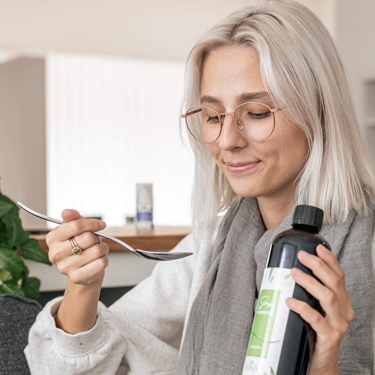 Frau hält Flasche und Löffel. Flasche mit Etikett. Frau trägt Brille und Schal. Löffel wird zum Mund geführt.