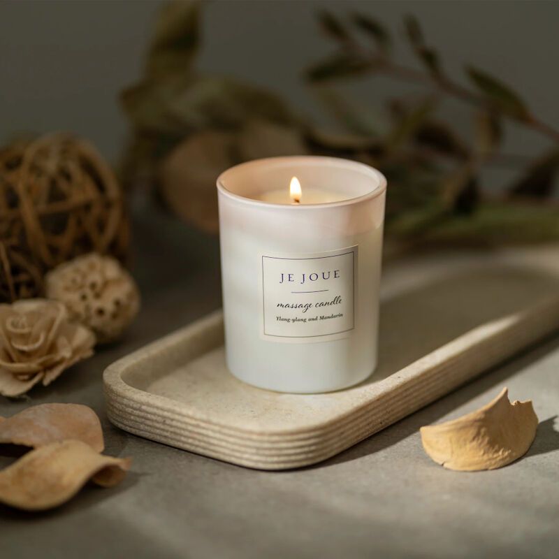 Brennende weiße Kerze in einem Glasbehälter. Auf einem Tablett. Label: Je Joue, massage candle. Hintergrund: Dekoration.