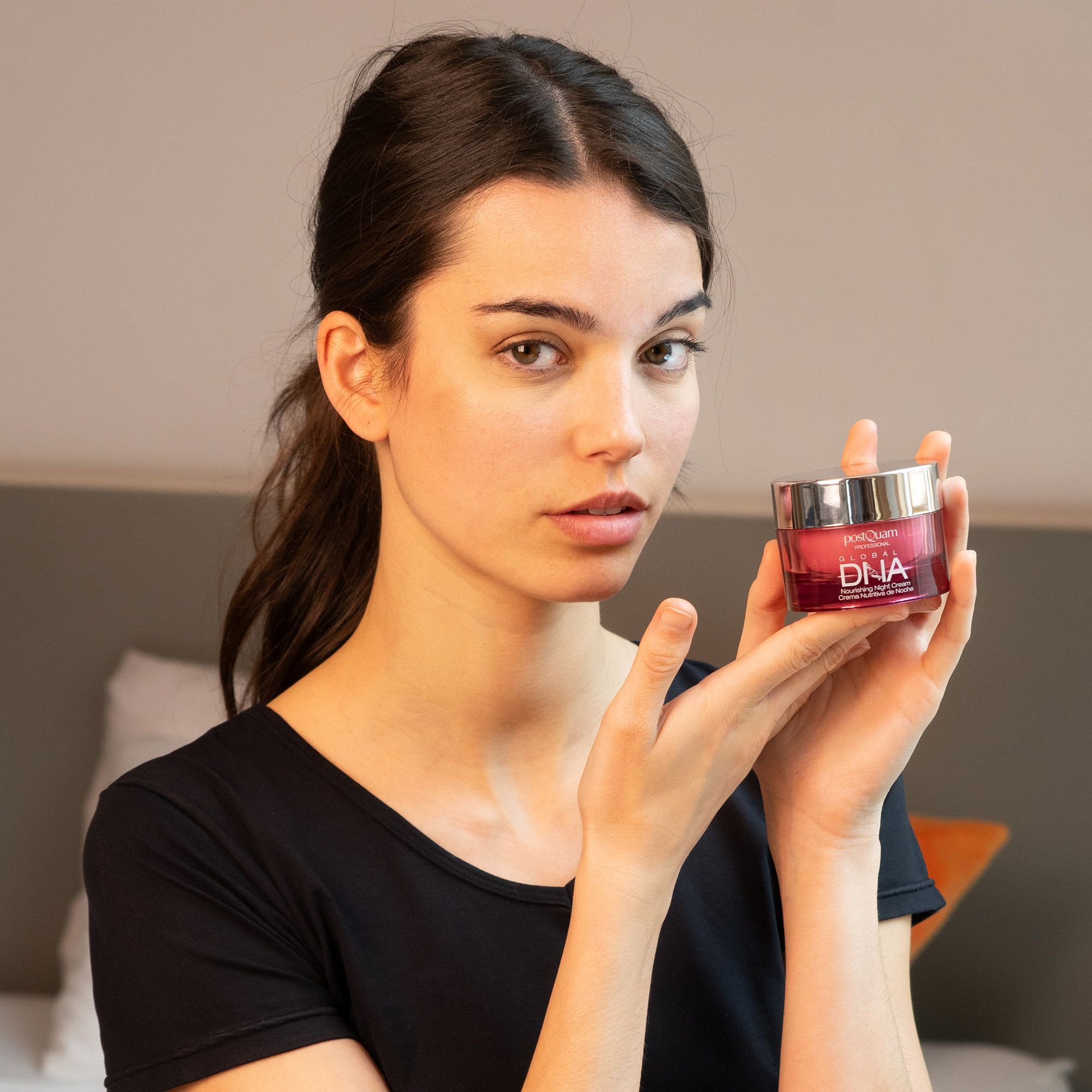 Frau hält Tiegel. Aufschrift: Postquam Global DNA Nourishing Night Cream. Rosa Tiegel mit silbernem Deckel, vor Person.