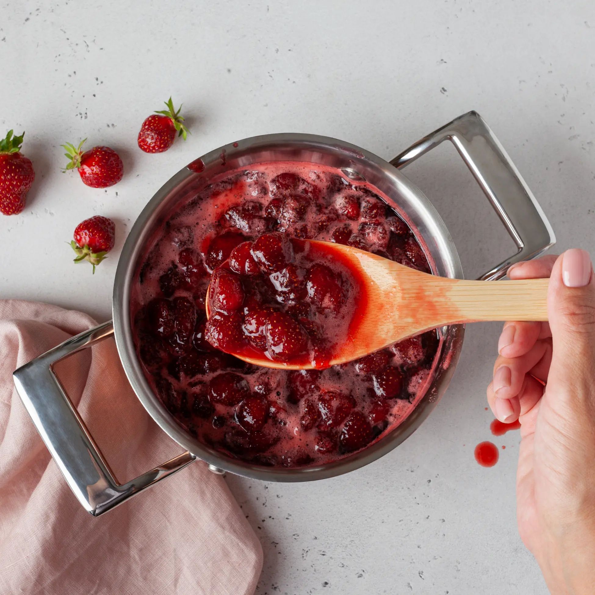 Topf mit Erdbeerkonfitüre. Ein Holzlöffel nimmt Konfitüre aus dem Topf. Daneben Erdbeeren.