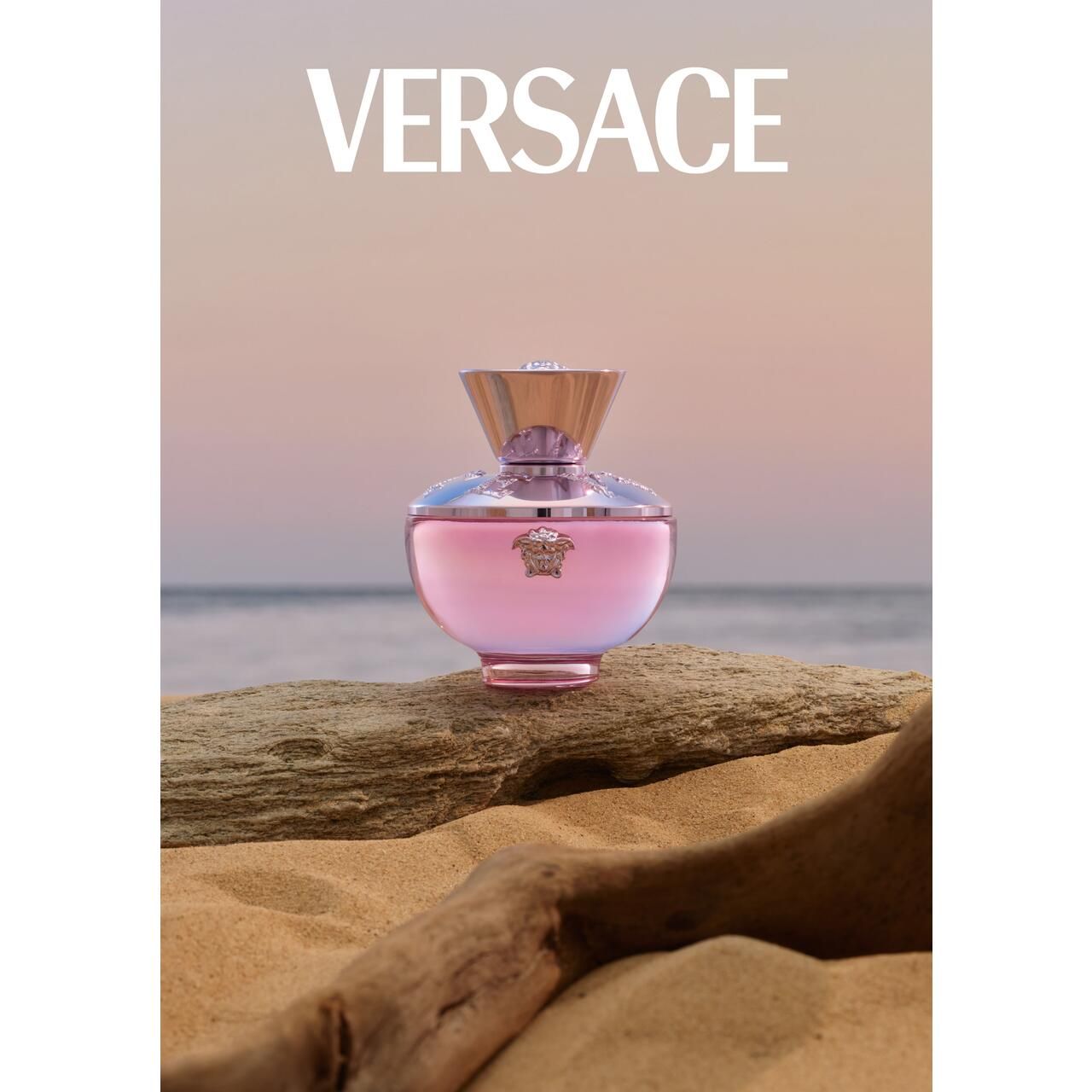 Versace Dylan Blush Pink Eau de Parfum Flakon auf Sand und Holz. Flakon mit rosa Glas, silbernem Deckel und Medusa-Logo. Hintergrund: Meer und Himmel.