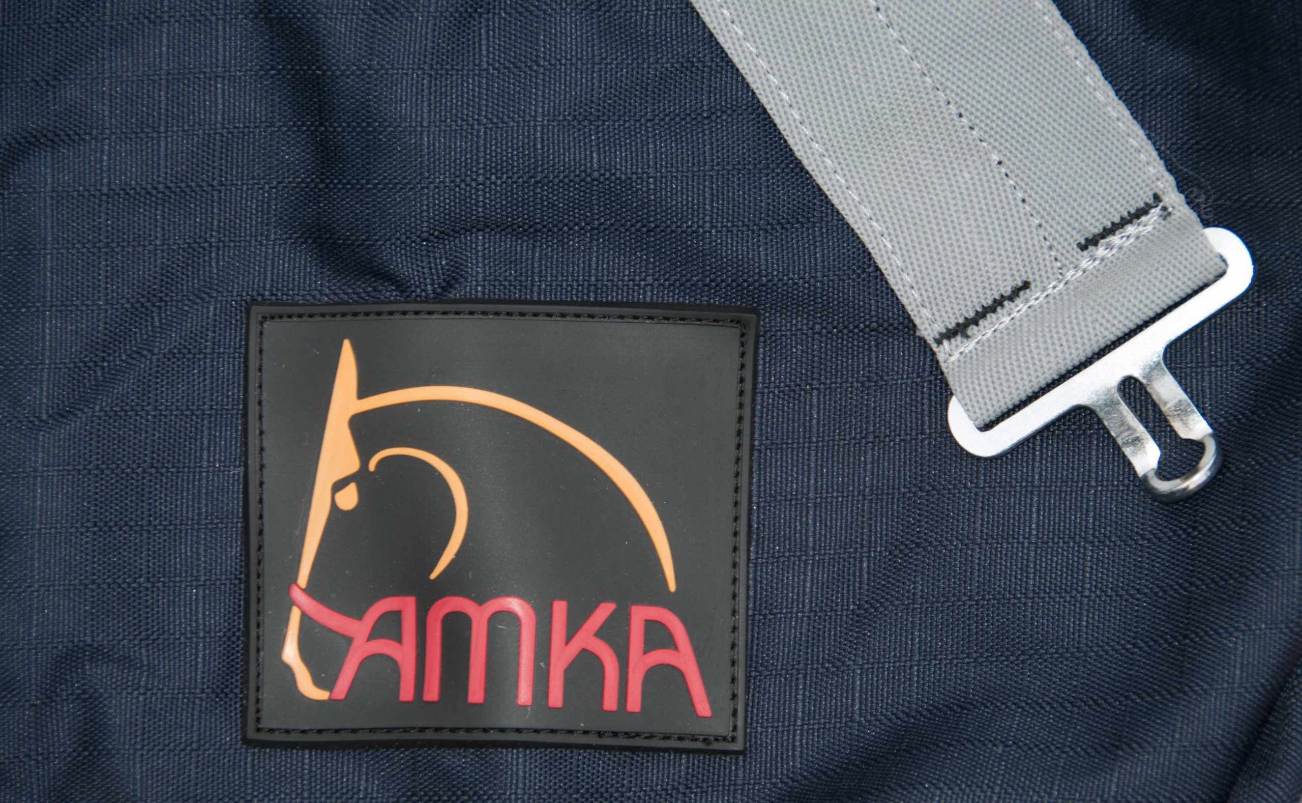Detailansicht des AMKA-Logos auf einem schwarzen Etikett. Das Logo zeigt einen Pferdekopf in Orange und Rosa.