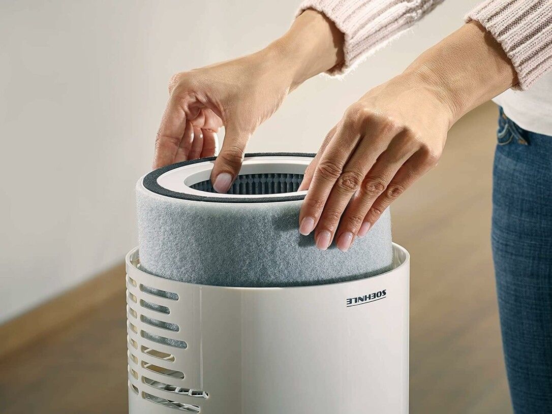 Hände setzen Filter in weißen Luftreiniger ein. Filter zylindrisch, grau. Luftreiniger mit Soehnle-Logo.