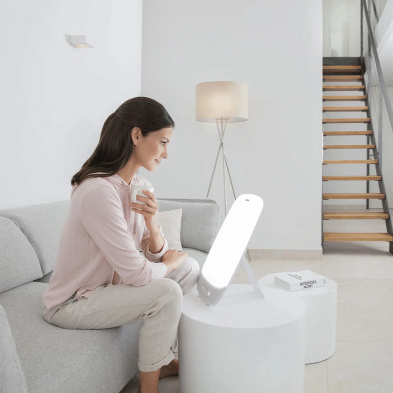 Frau sitzt auf Sofa, vor weißer Tageslichtlampe. Auf Tisch, Buch und Lampe.
