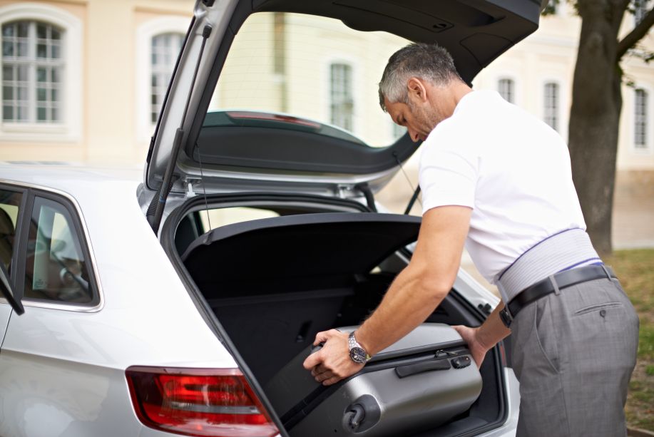 Mann lädt Koffer in Auto ein, trägt LordoLoc-Rückenbandage. Graue Bandage unter Hemd sichtbar. Auto im Hintergrund.