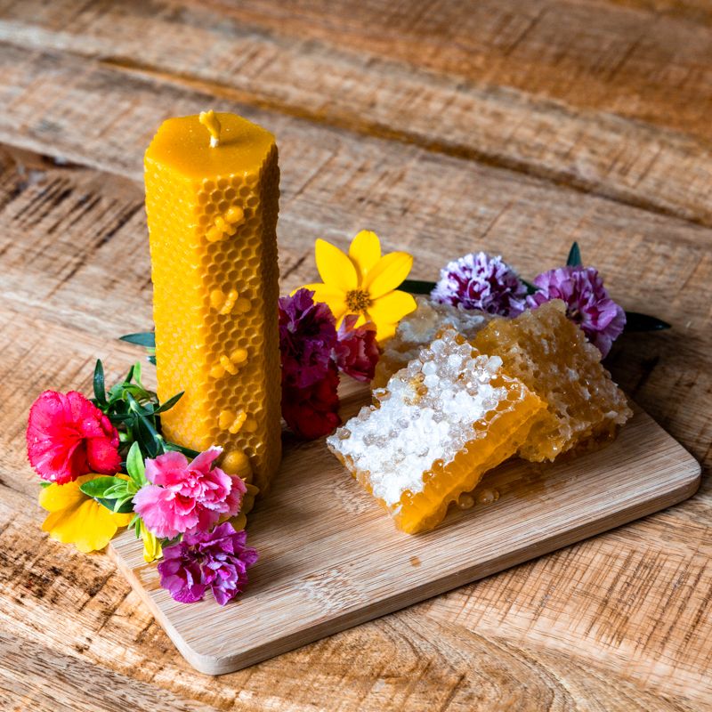 Gelbe Wachskerze mit Bienenwabenmuster, brennend. Neben Honigwaben und Blumen auf Holzbrett.
