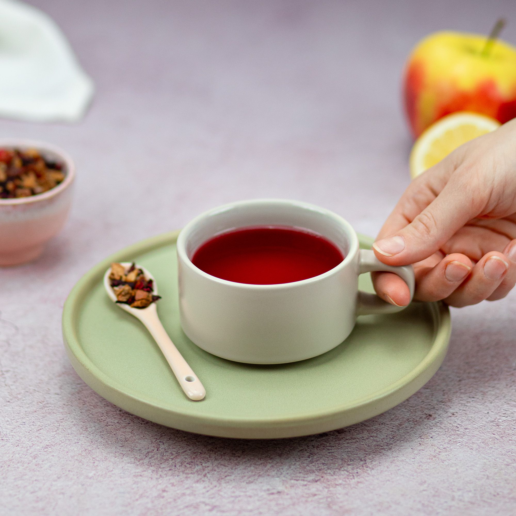 Tasse Tee auf Untertasse. Löffel mit Teemischung. Hand hält Tasse. Apfel und Zitrone im Hintergrund.