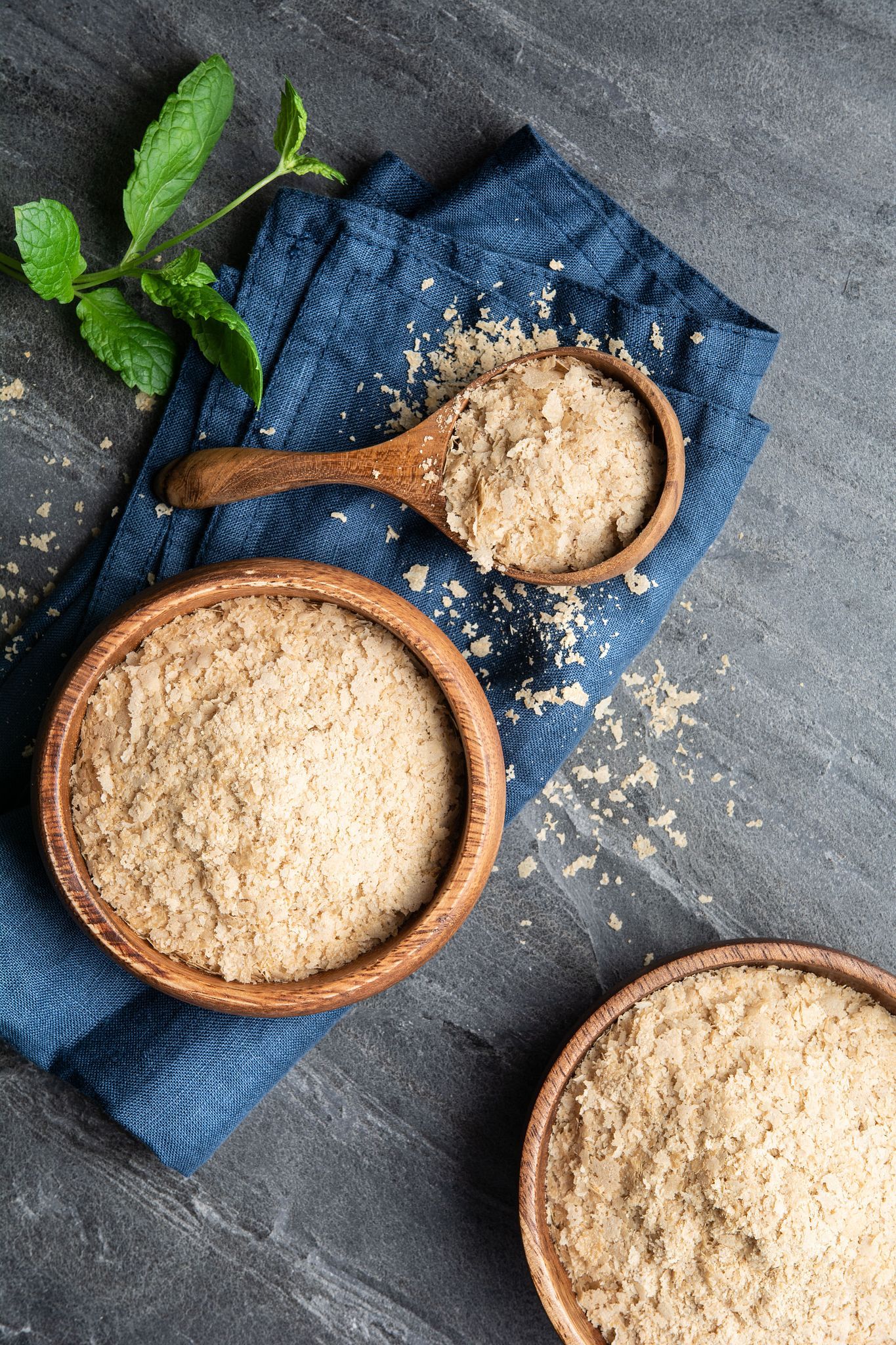 Drei Holzschalen mit Hefeflocken. Eine Schale mit Löffel auf blauem Tuch. Minzzweig.