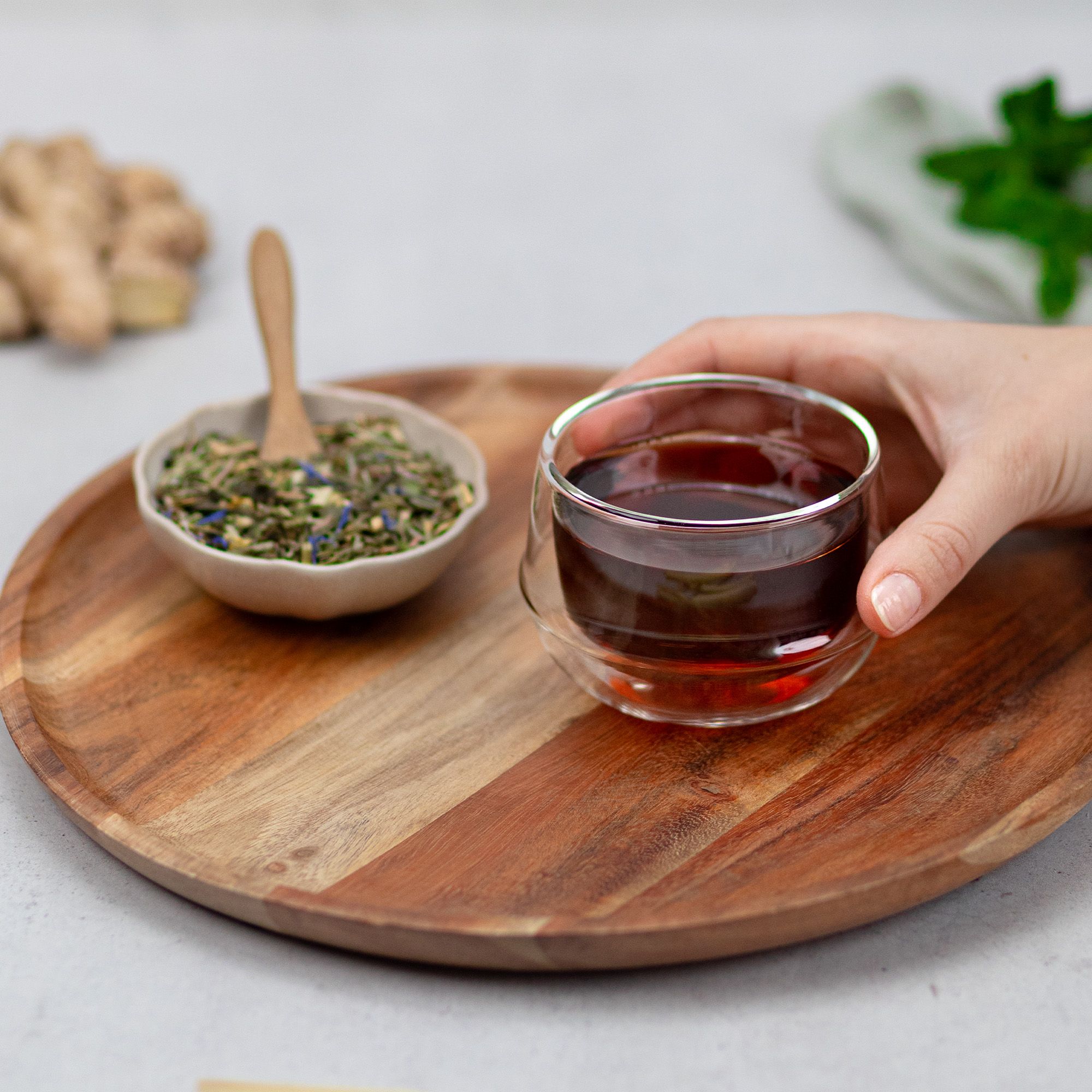 Glas mit rotem Tee auf Holztablett. Daneben Schale mit Kräutern und Holzlöffel. Hintergrund unscharf.