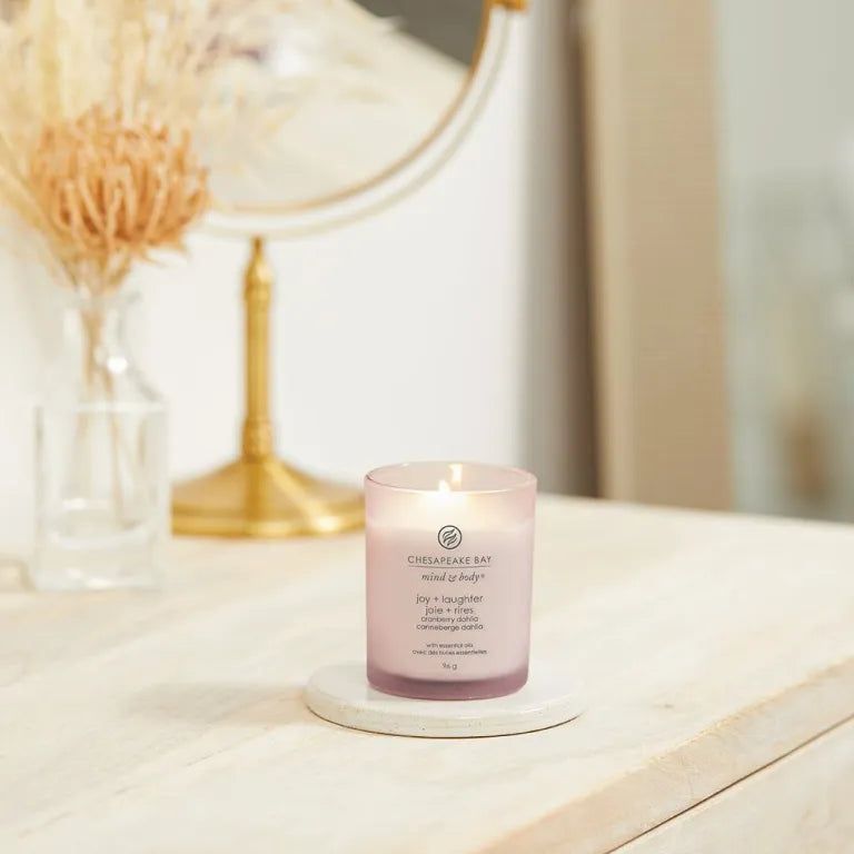 Rosa Kerze Chesapeake Bay mind & body, joy + laughter, cranberry dahlia, auf Untersetzer. Spiegel und Vase im Hintergrund.