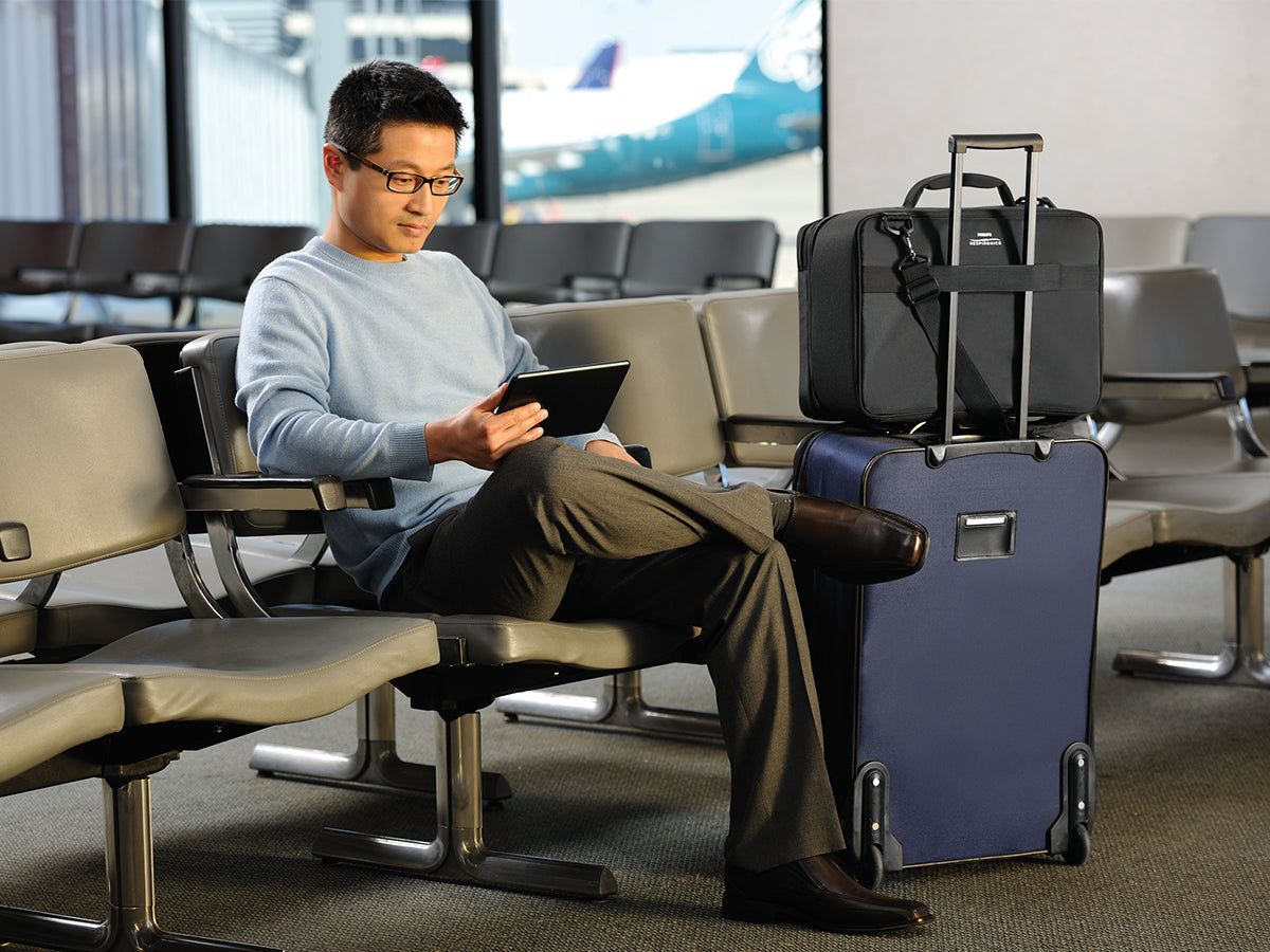 Mann sitzt in Flughafen-Wartebereich mit schwarzer Reisetasche auf einem Rollkoffer neben sich.