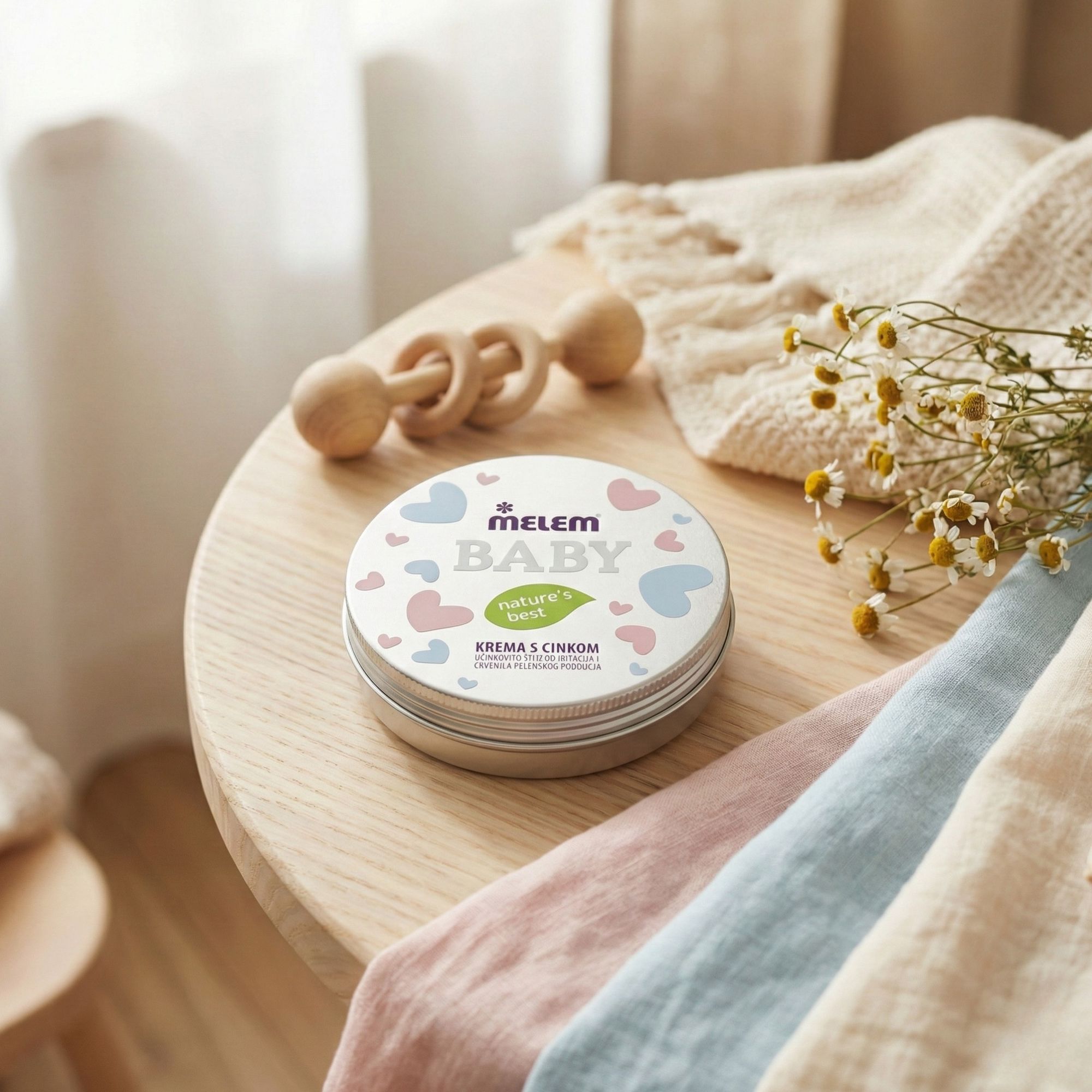 Runde Dose mit Aufschrift Melem Baby auf einem Tisch. Daneben ein Holzrassel und Stoffwindeln.