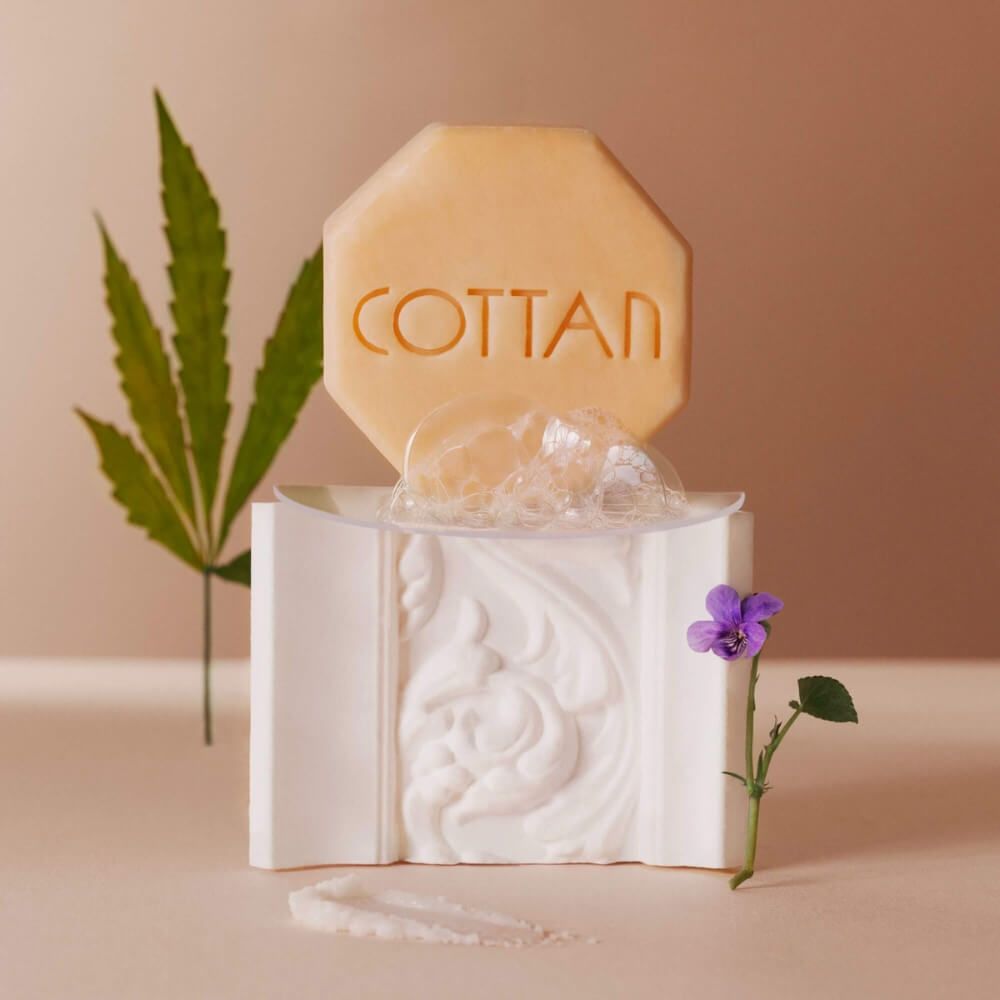 Achteckige Seife mit COTTAN-Schriftzug, auf weißem Podest mit Blasen. Daneben ein Blatt und eine Blume.