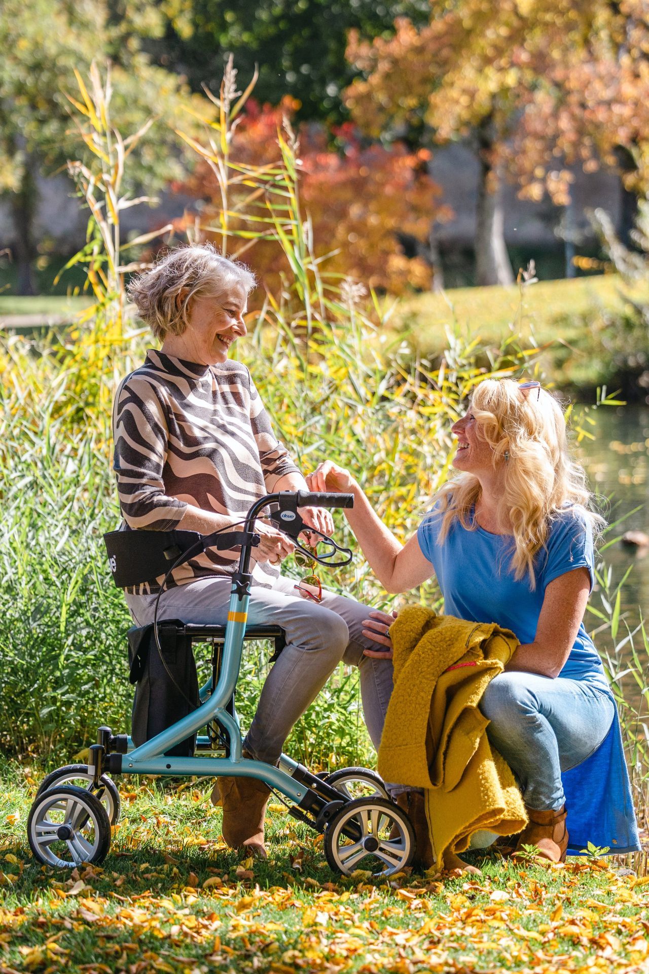 Rollator, blau, mit zwei Frauen im Park. Eine sitzt, die andere steht daneben. Herbstliche Umgebung.